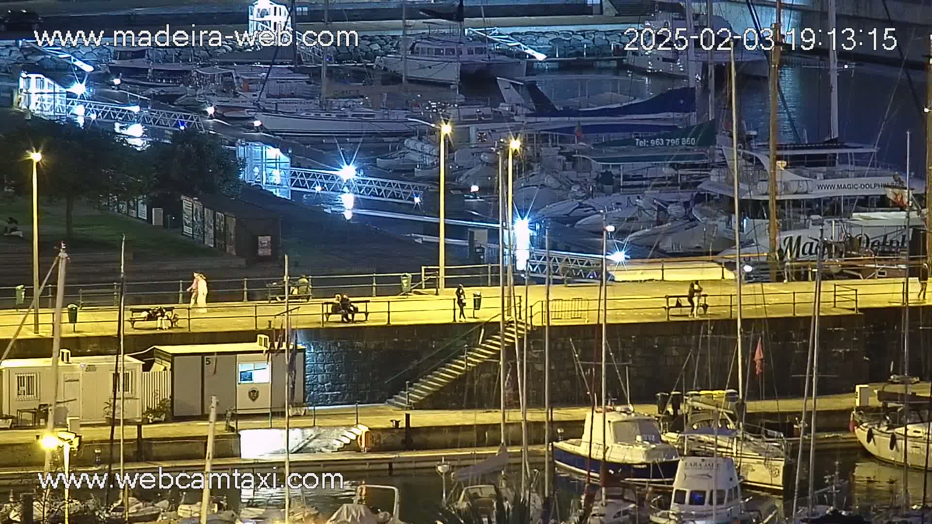 A nighttime view of a marina, with numerous boats docked, and several people walking along a lit waterfront walkway.