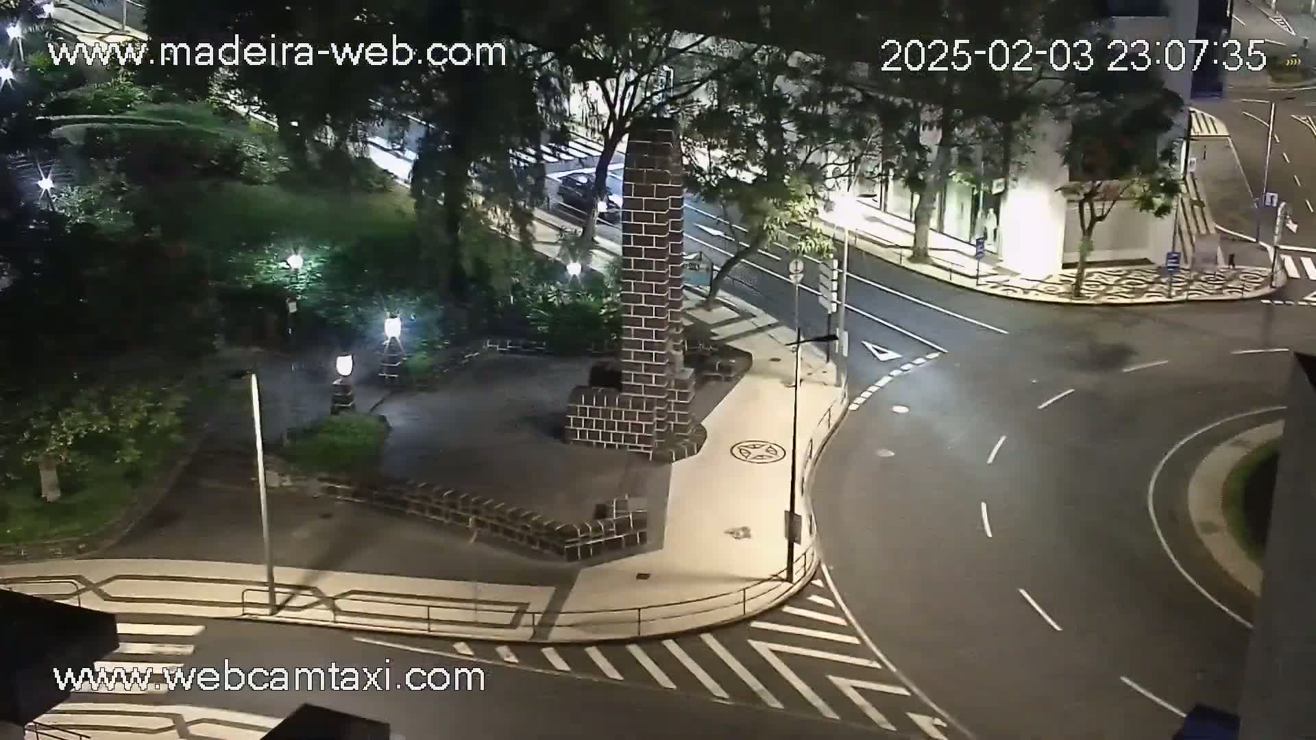 A nighttime, high-angle view shows a brick monument in a small plaza next to a curved road intersection with sparse traffic under partly cloudy skies.