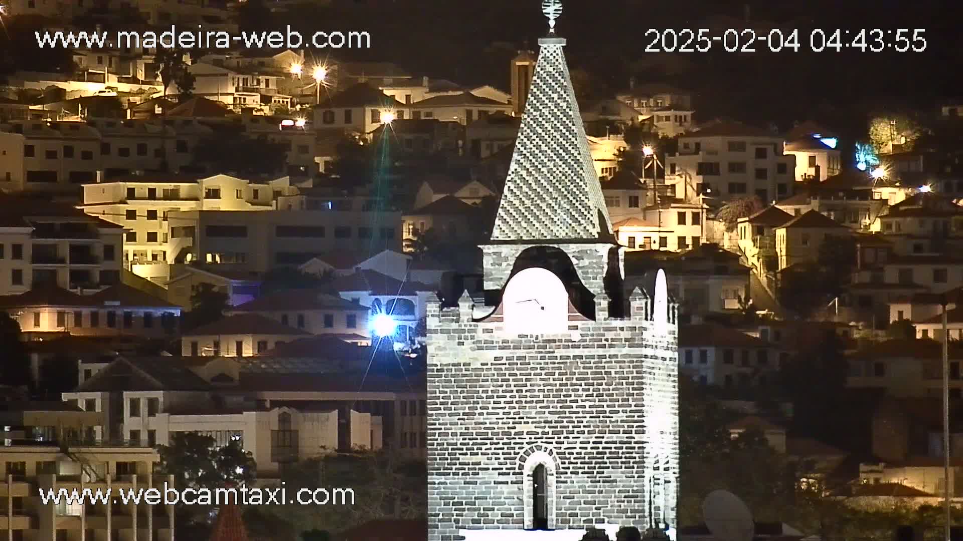 A brick tower with a pointed top stands prominently against a backdrop of a nighttime hillside town illuminated by streetlights.