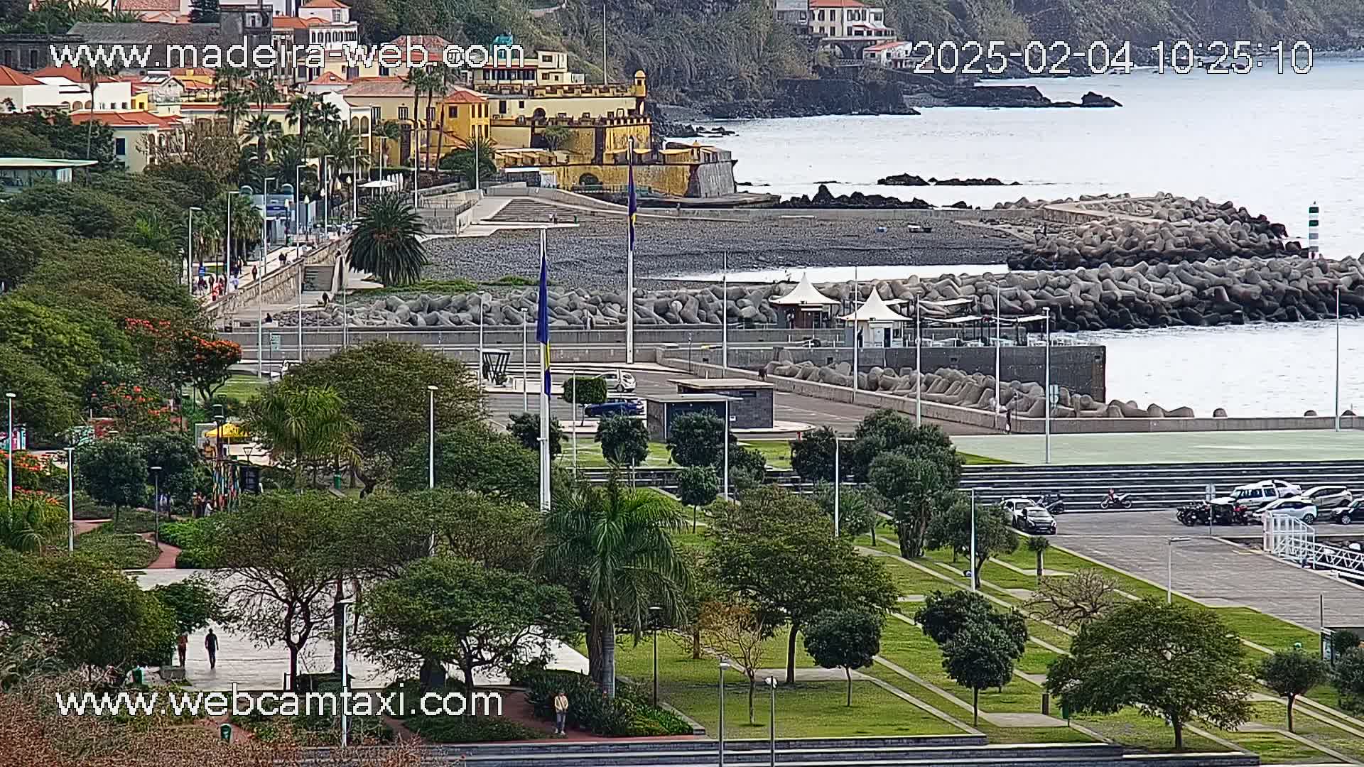 A coastal town with a park, a paved walkway, and a rocky shoreline, under overcast skies.