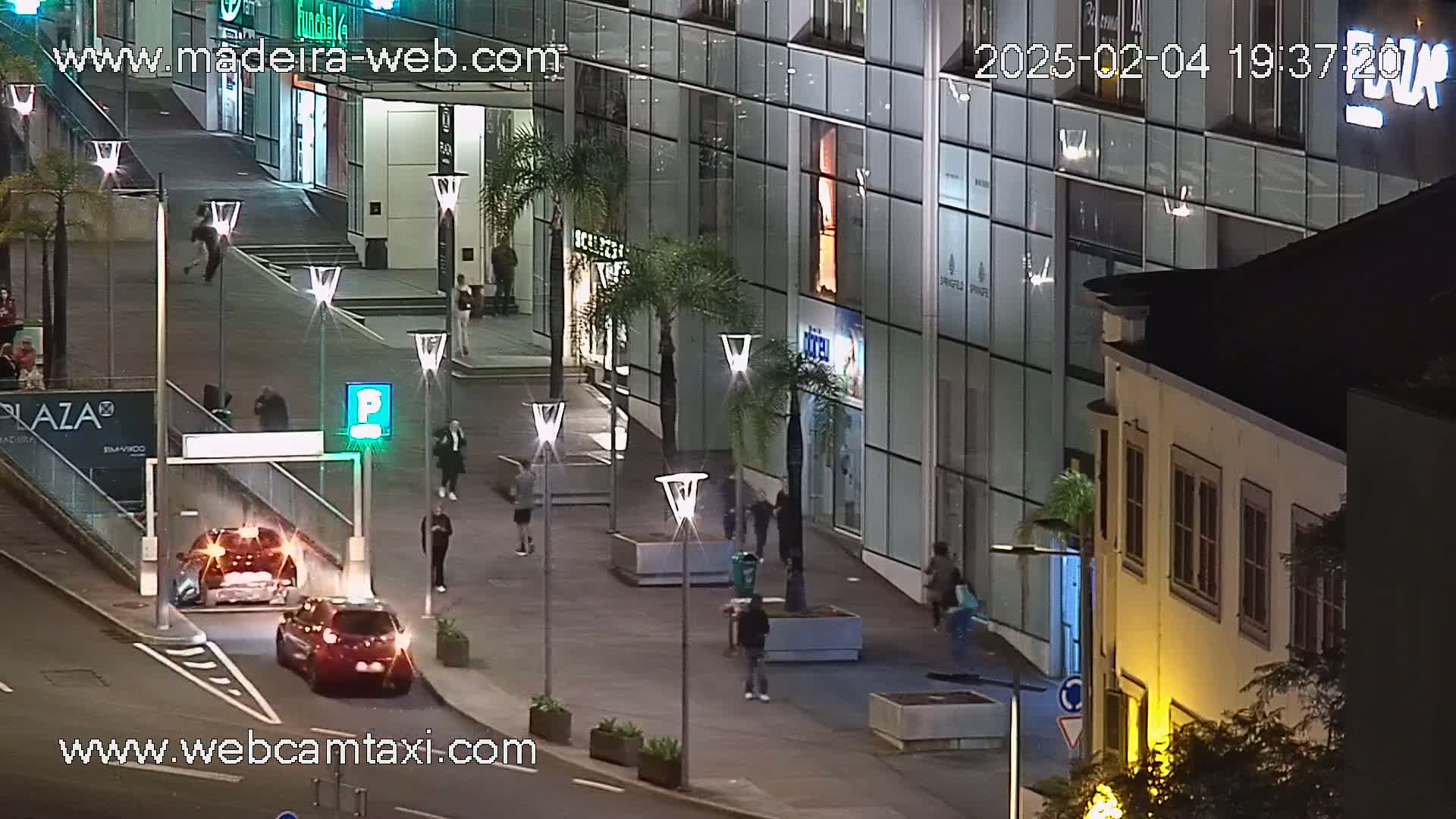 At night, several people walk along a city sidewalk in front of shops and buildings, with two cars visible on a street nearby.