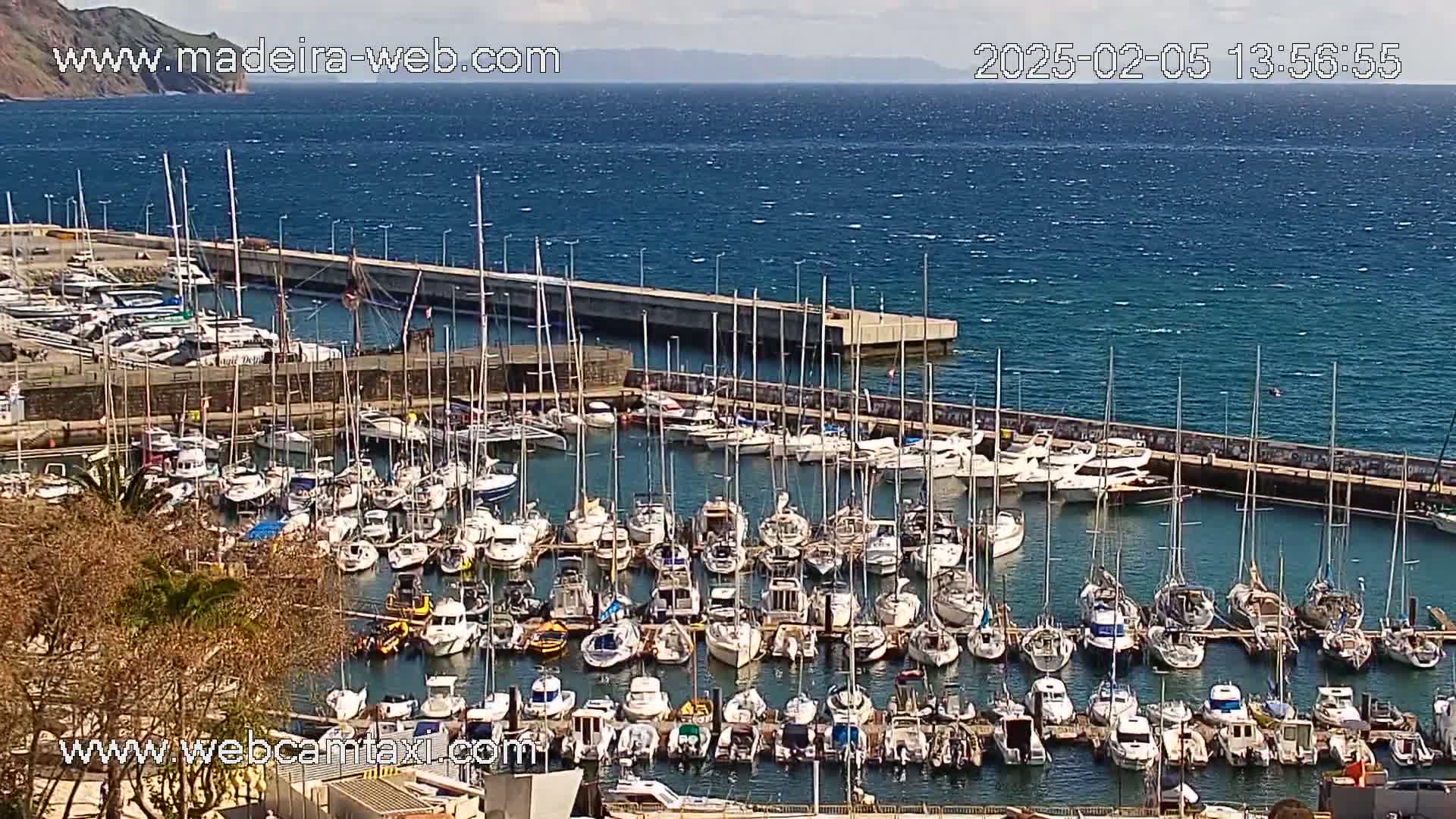 A marina filled with numerous sailboats and motorboats is seen from an elevated position on a partly sunny day with a somewhat windy ocean in the background.