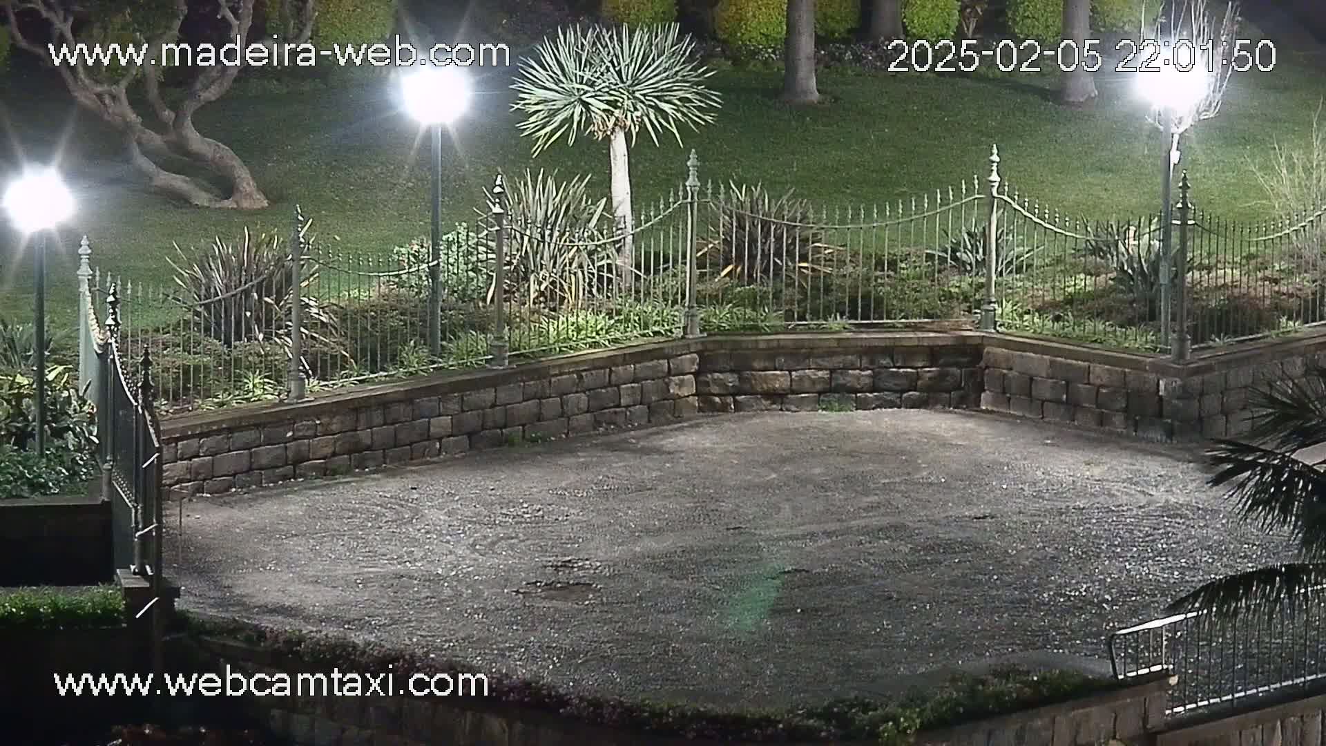 A nighttime view of an empty, paved area enclosed by a low stone wall and wrought-iron fence, illuminated by several streetlights, surrounded by lush green landscaping.