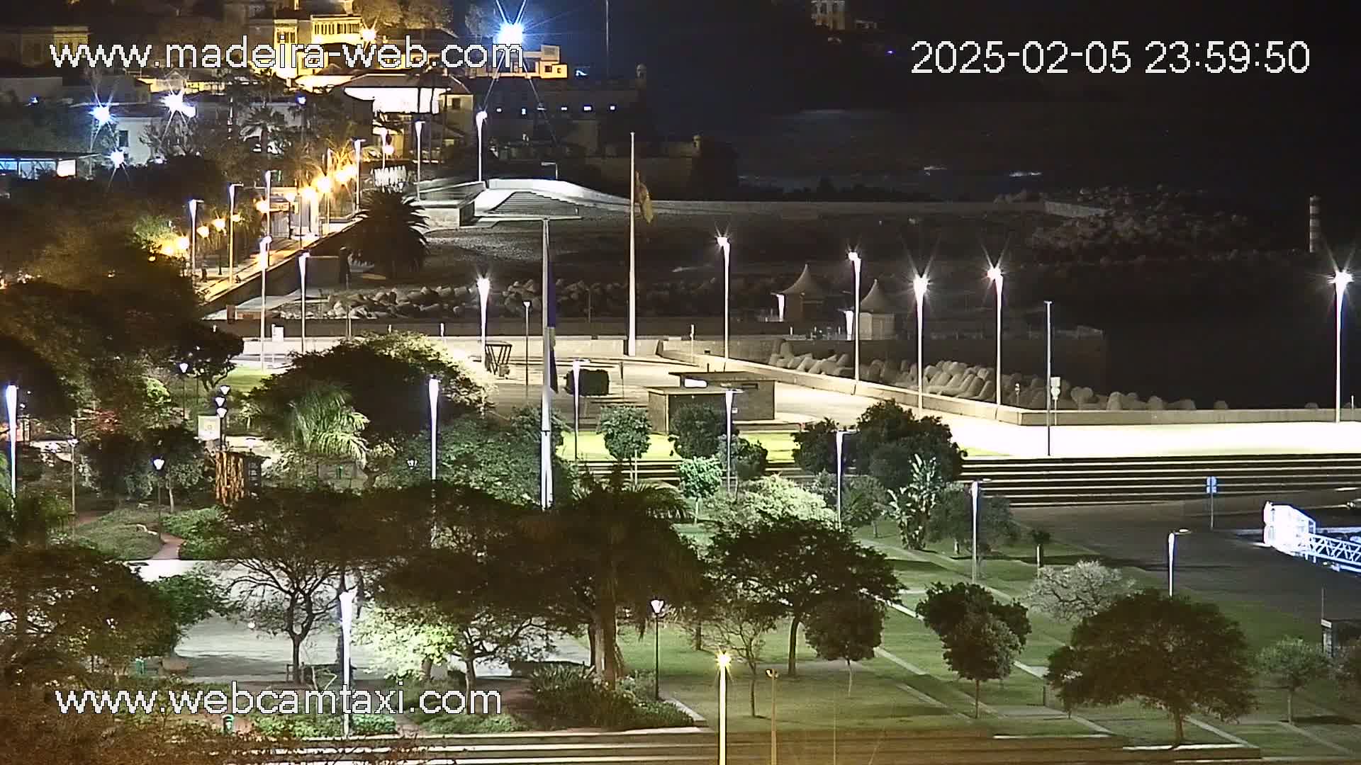 A nighttime view of a park and coastal area, illuminated by streetlights, with a calm ocean in the background.
