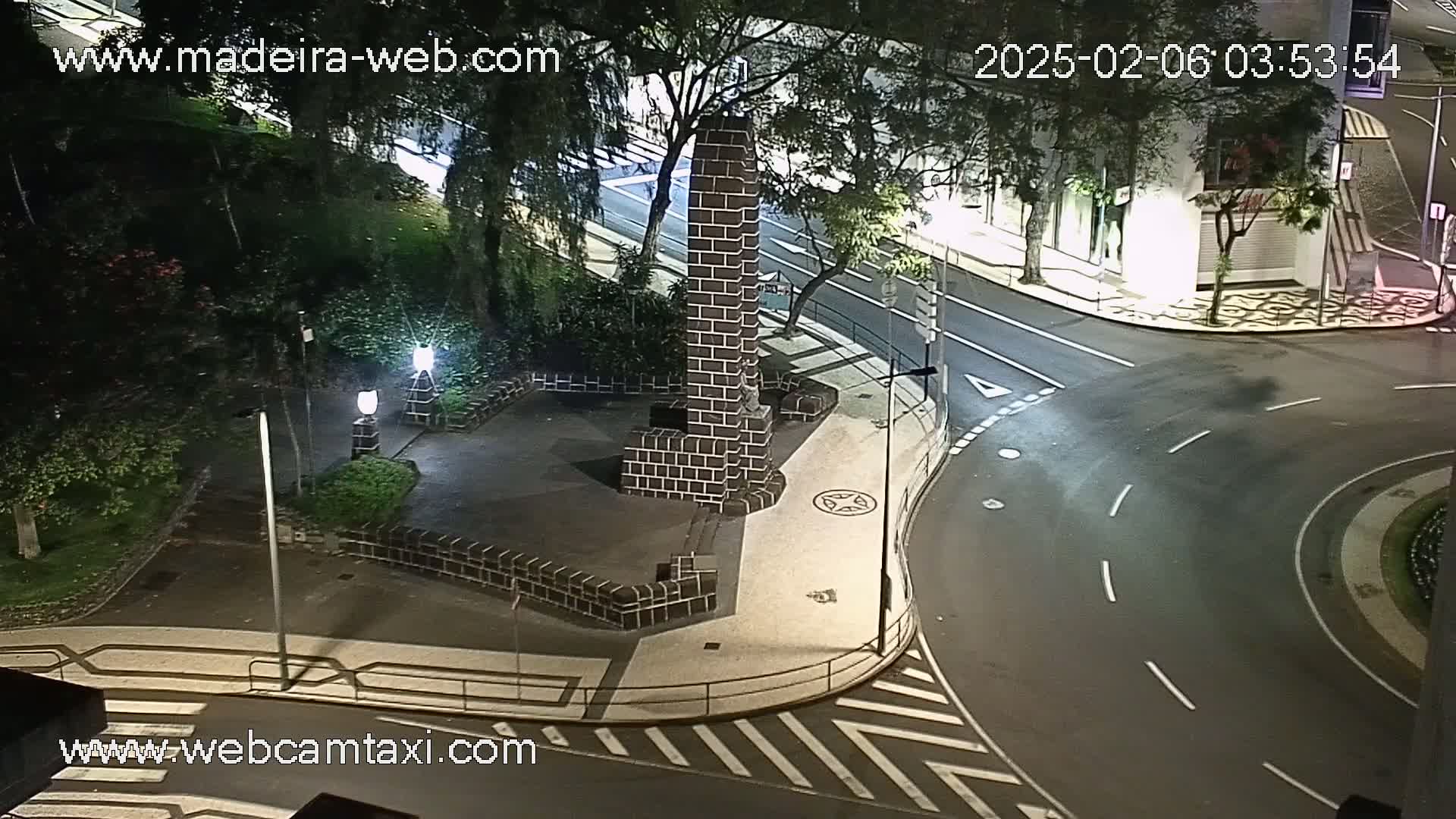 A nighttime, high-angle view of a street intersection with a dark brick monument in a small plaza, surrounded by trees and streetlights.