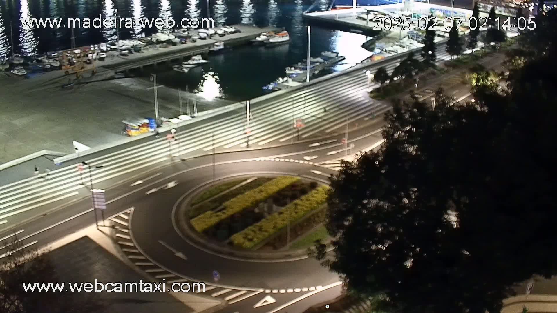 A nighttime aerial view of a harbor with boats docked, a roundabout with landscaping in the center, and a roadway leading to a staircase that overlooks the harbor.