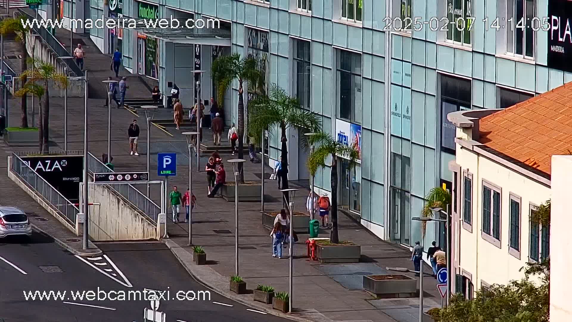 A sunny day shows pedestrians walking on a city sidewalk in front of modern glass buildings and older, yellow buildings with orange tile roofs, near palm trees.