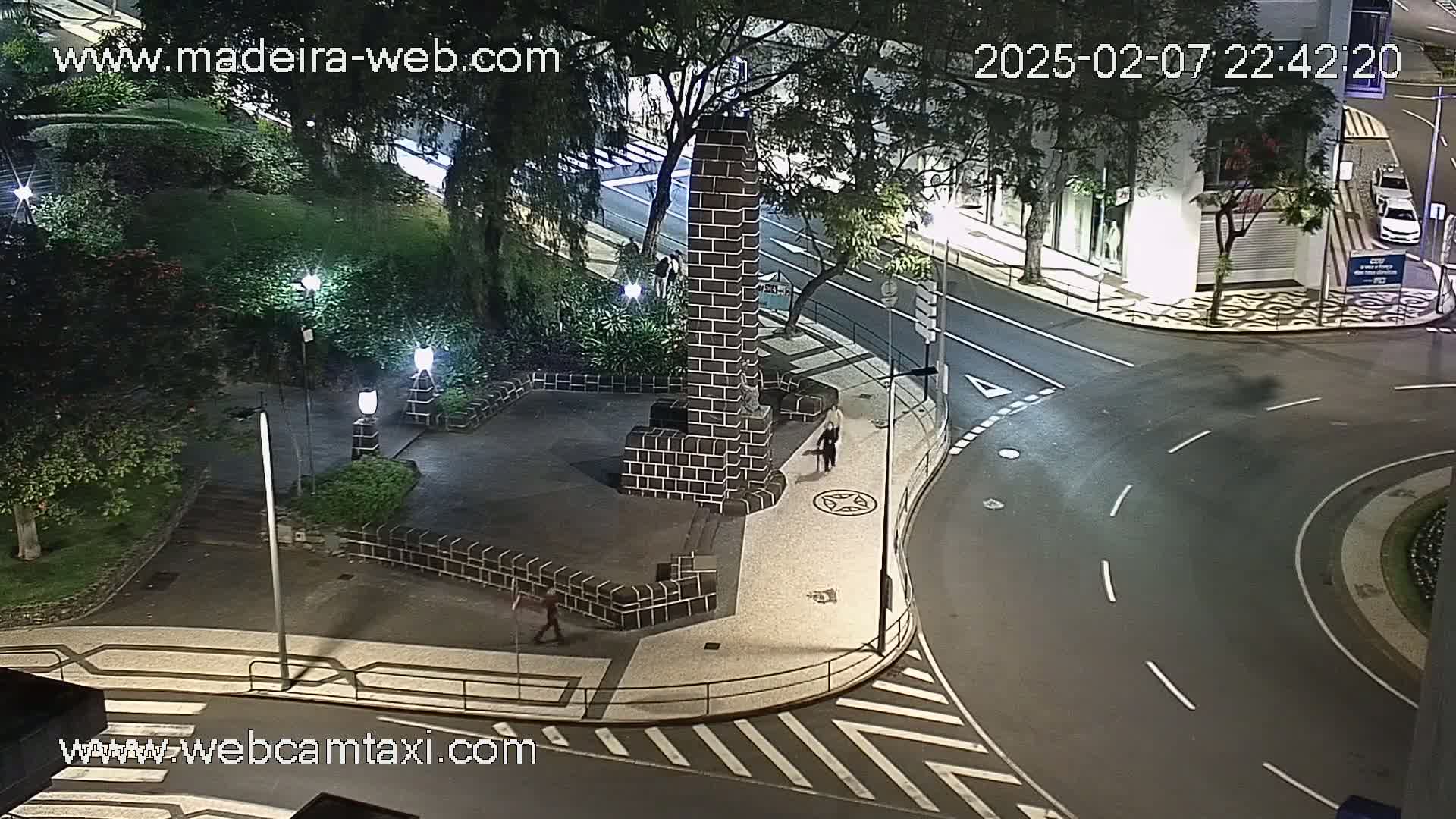 A nighttime view of a paved plaza with a dark brick monument, surrounded by trees and streetlights, near a curved road.