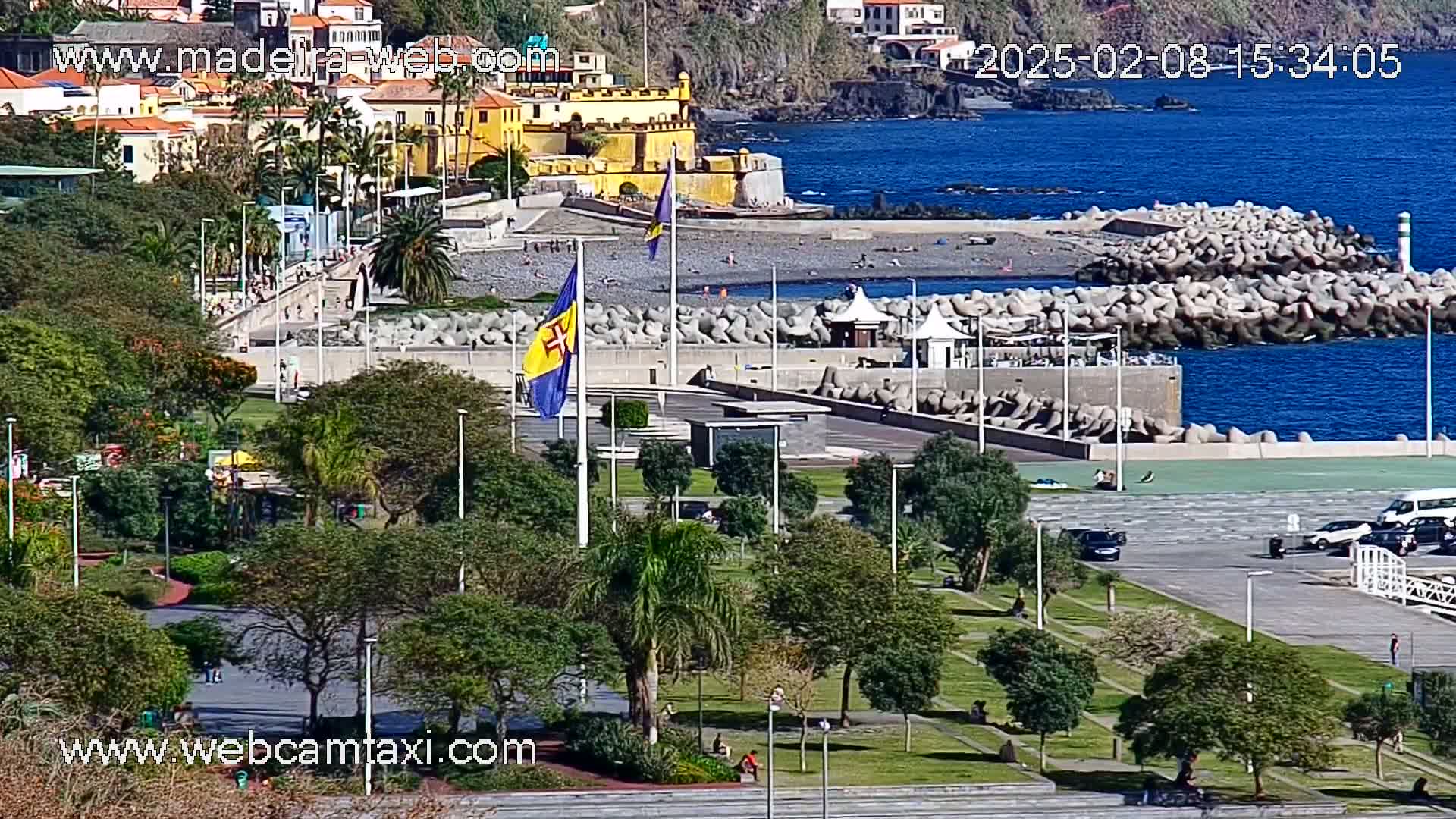 A sunny coastal scene shows a park with lush greenery next to a paved area, a pebble beach, a breakwater, and a yellow fort overlooking the calm blue ocean.