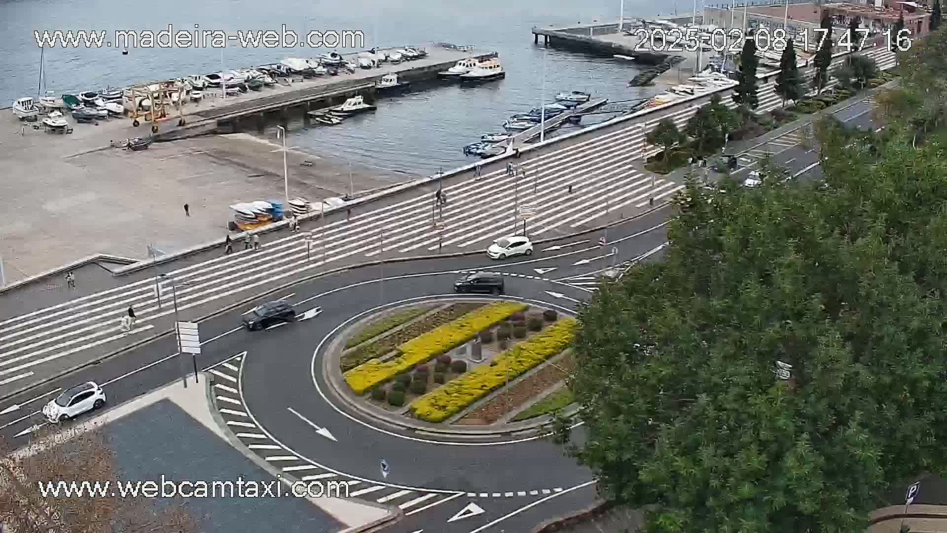 An aerial view shows a harbor with boats, a roadway with cars and pedestrians, and a roundabout with a landscaped center, all under overcast skies.