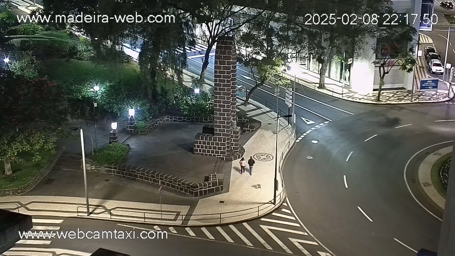 A nighttime, high-angle view shows a dark brick monument in a plaza next to a curved roadway with two pedestrians walking nearby.
