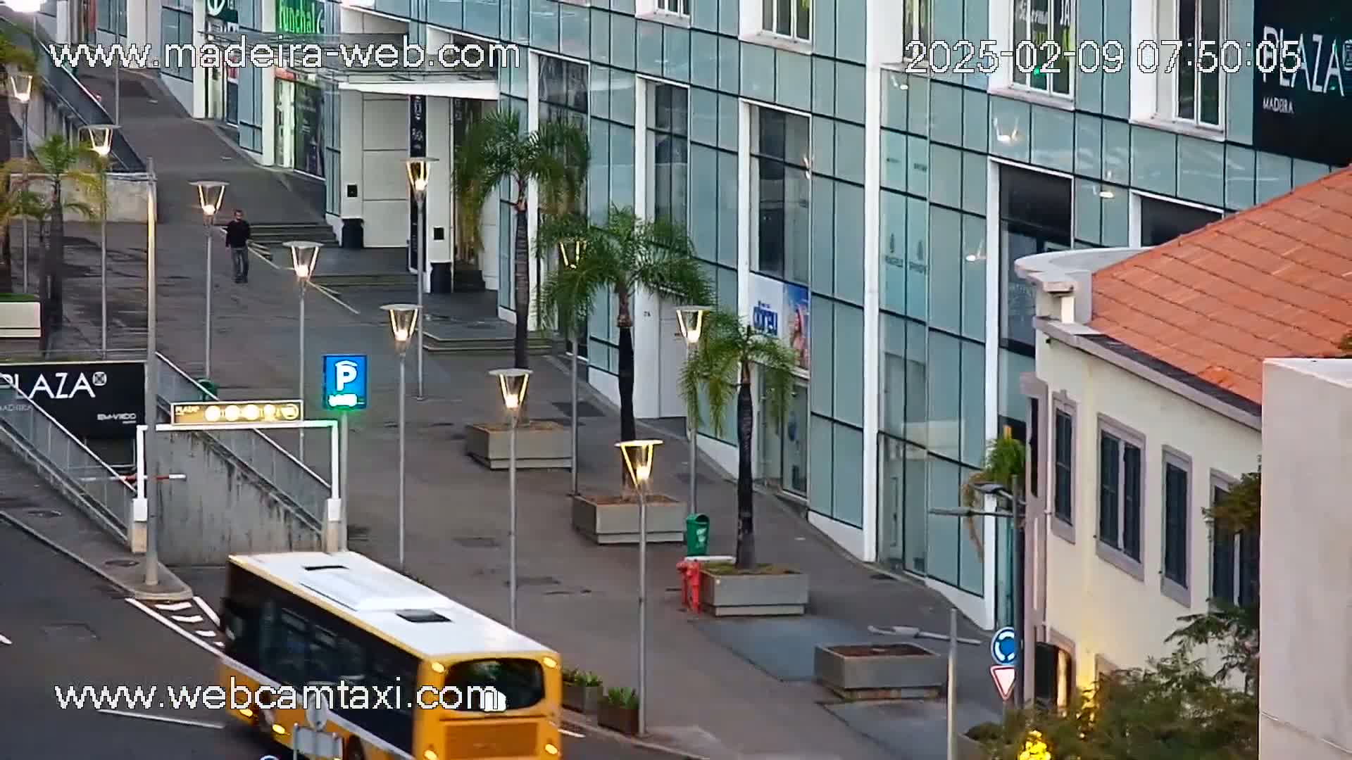 A yellow bus drives past a modern glass building and a smaller, older building with a red tile roof on a partly sunny day.