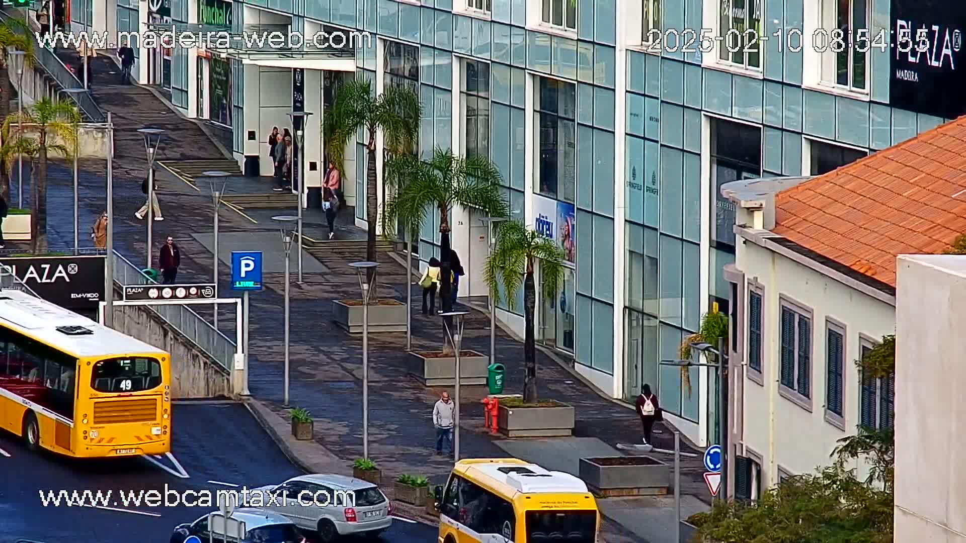 A wet street scene shows a yellow bus and several pedestrians near a modern glass building and an older, smaller building with a red tile roof.