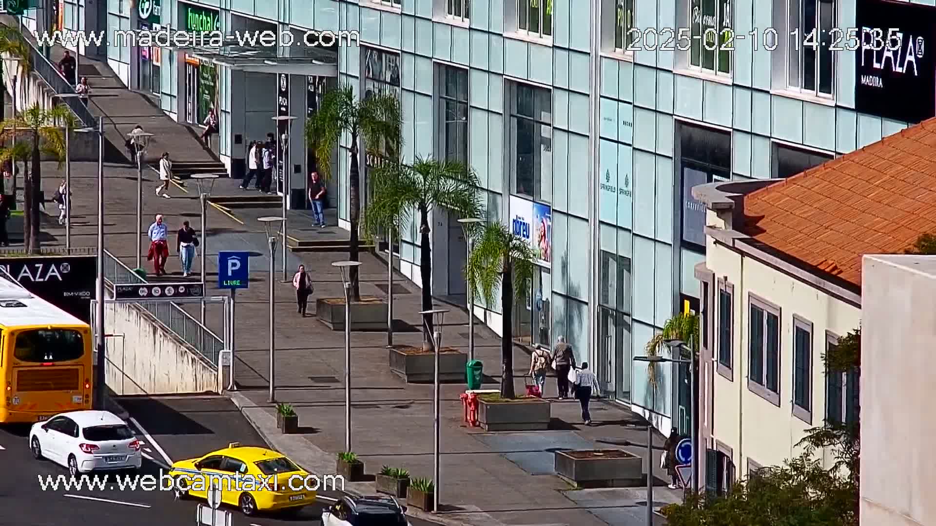 A sunny city street scene shows pedestrians walking past a modern glass building and a smaller, older building with a tiled roof, near a bus and cars.