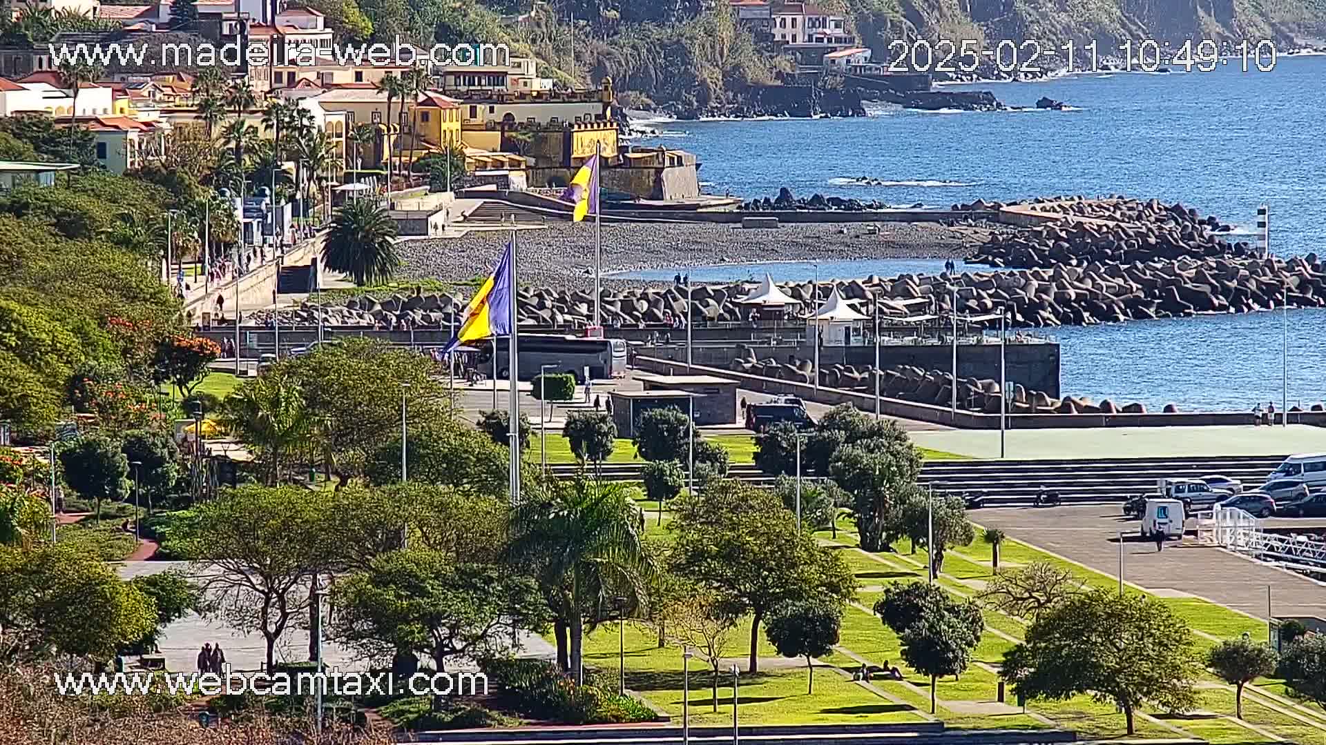 A sunny coastal scene shows a paved area with green lawns, trees, and a few people, leading to a rocky breakwater and a calm ocean with buildings and a flag in the background.