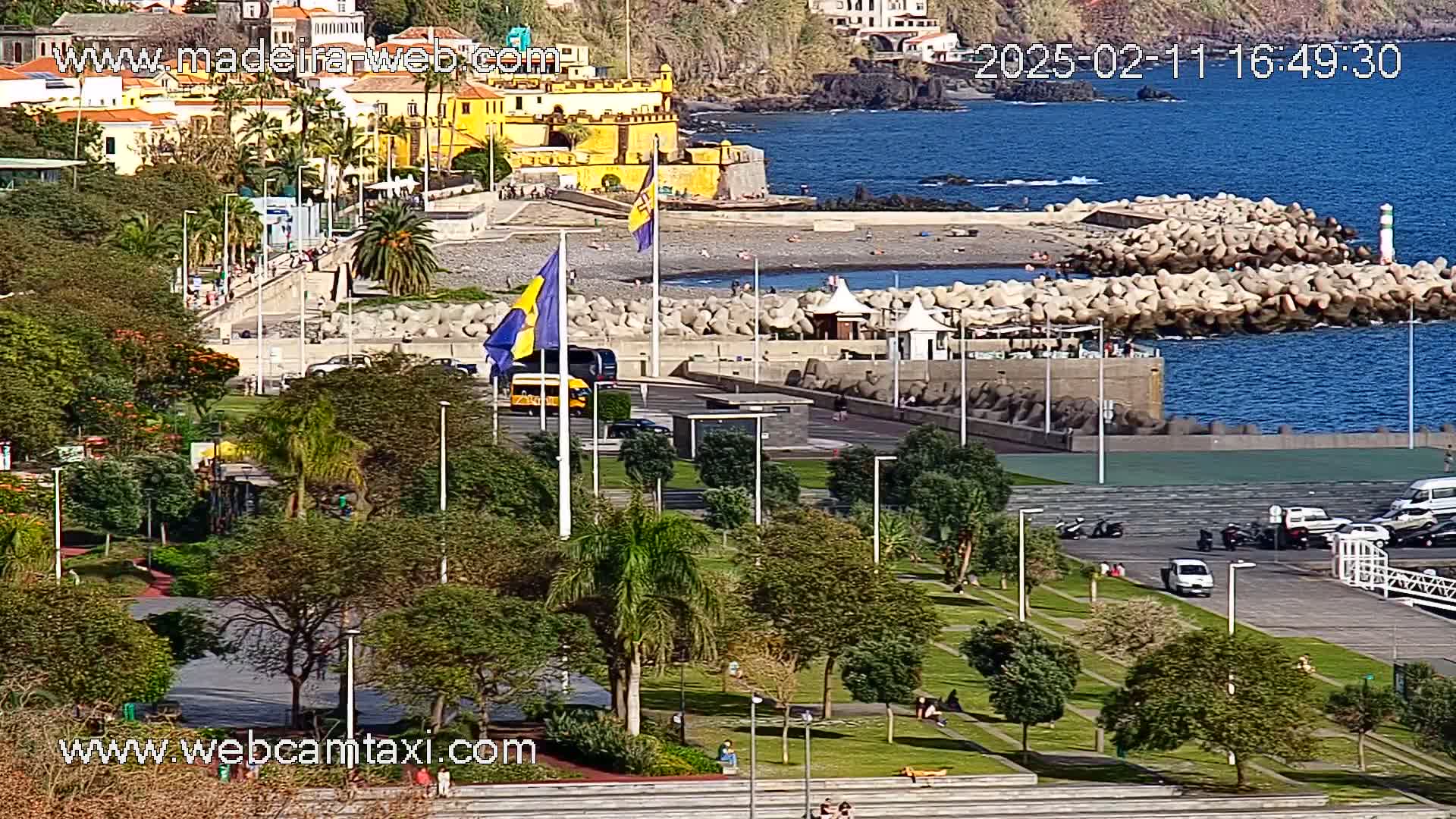 A sunny coastal scene shows a park with green lawns, palm trees, and people, overlooking a paved area with parked cars near a rocky shoreline and a yellow fortress.