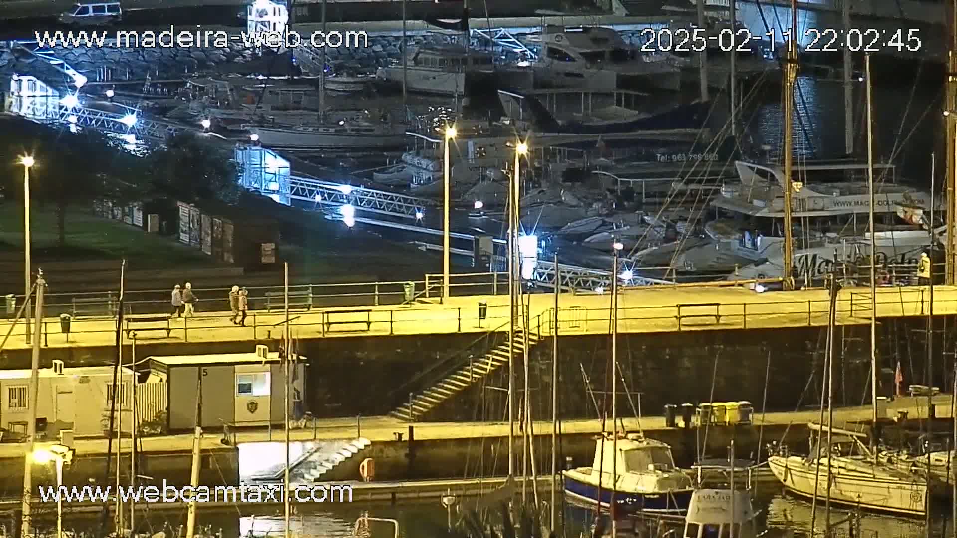 A nighttime view of a marina with numerous boats docked, several people walking along a waterfront walkway, and some small structures near the water's edge.