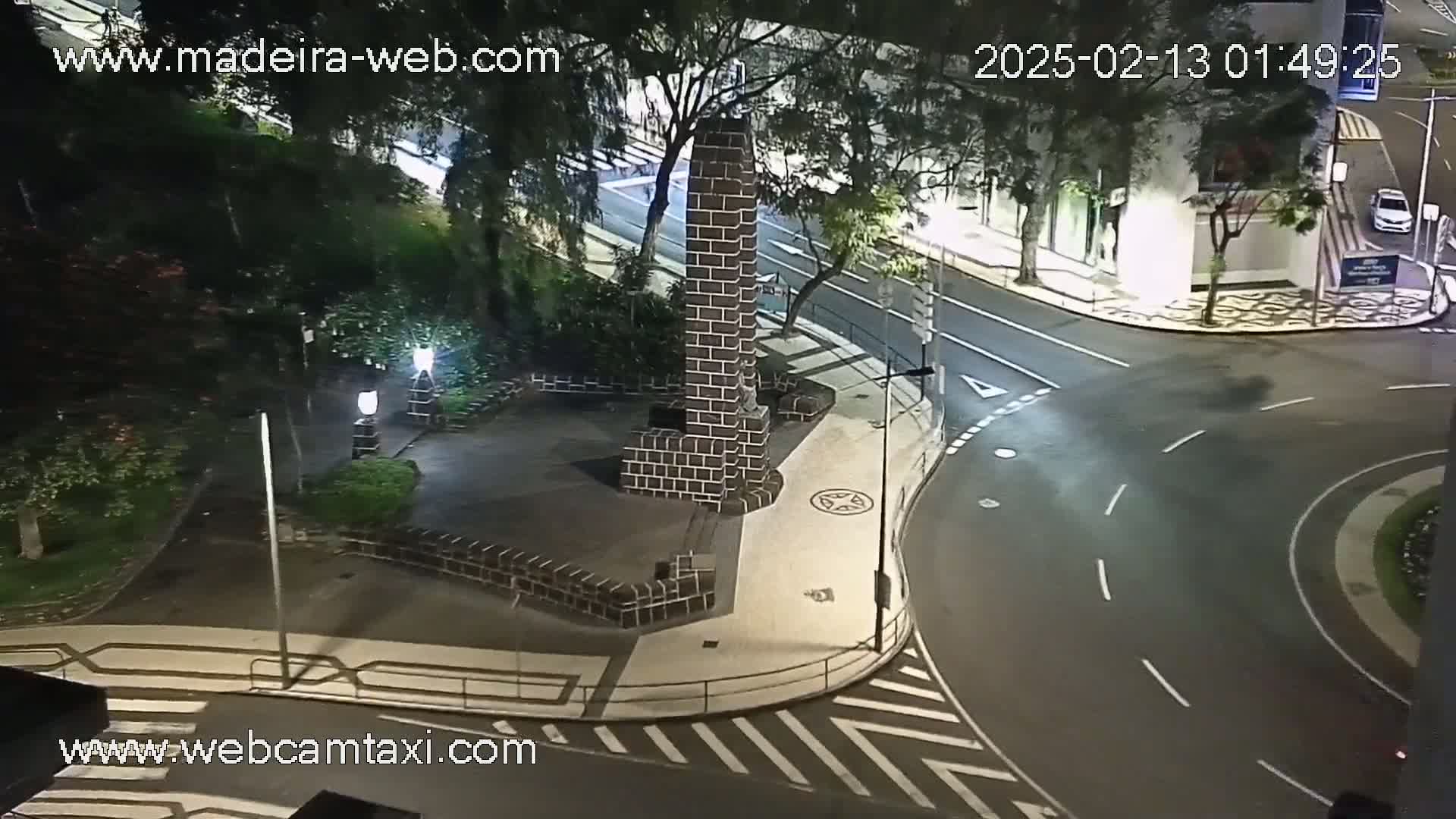 A nighttime overhead view of a paved intersection with a brick monument in a small plaza, surrounded by trees and streetlights.