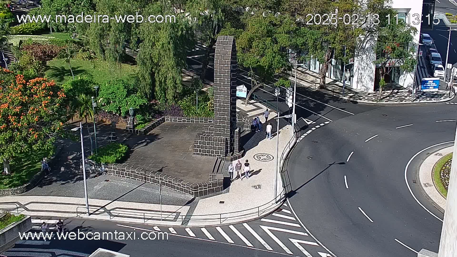 A sunny day shows a paved plaza with a dark stone monument, surrounded by lush green landscaping and a curved roadway.