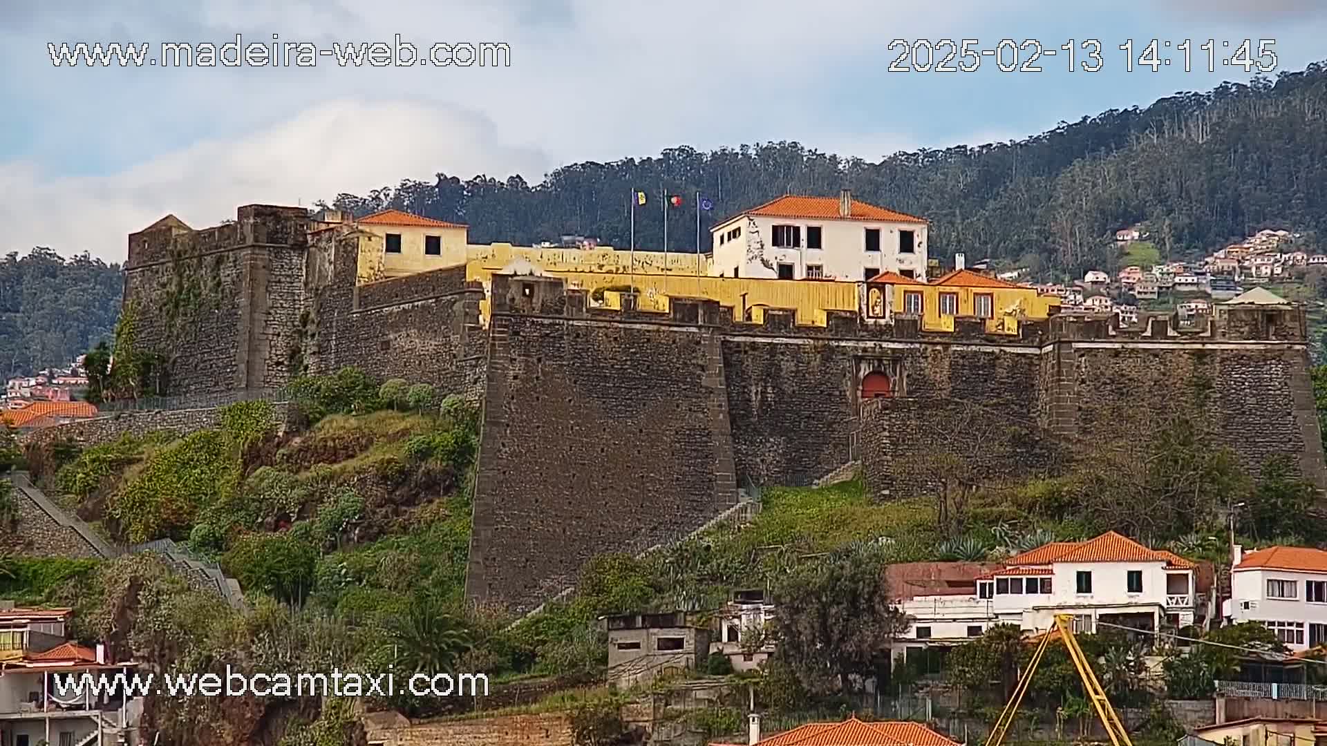 A stone fortress sits atop a hill overlooking a town under a partly cloudy sky.
