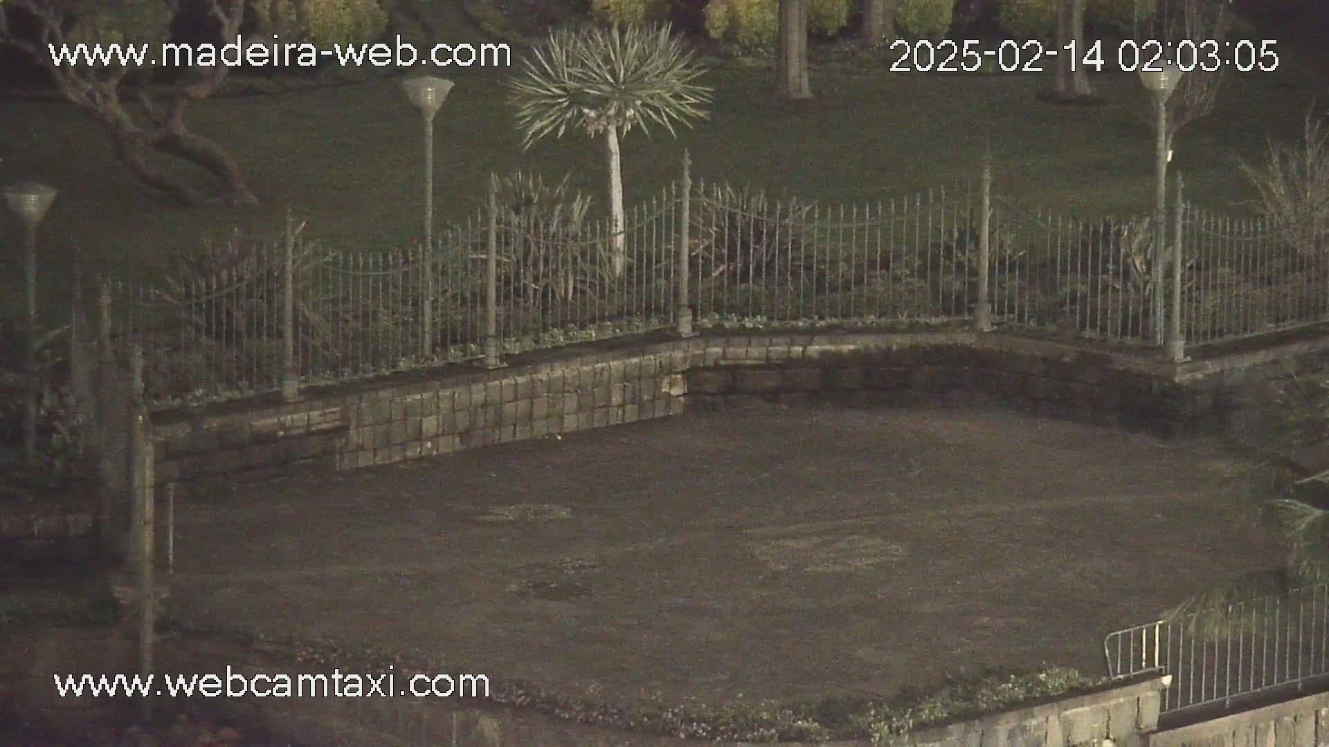 A nighttime, low-light view shows a dark paved area enclosed by a low stone wall and a metal fence, with some sparse vegetation visible beyond the fence.