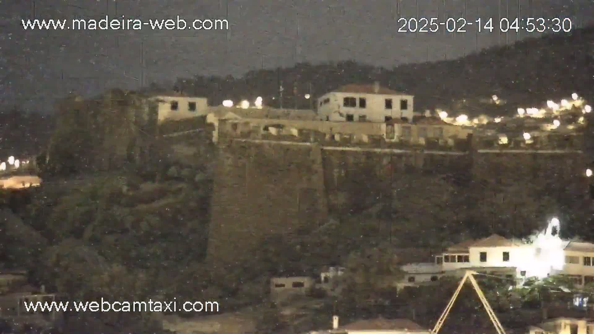 A nighttime view of a stone fortress and surrounding buildings under snowy conditions.