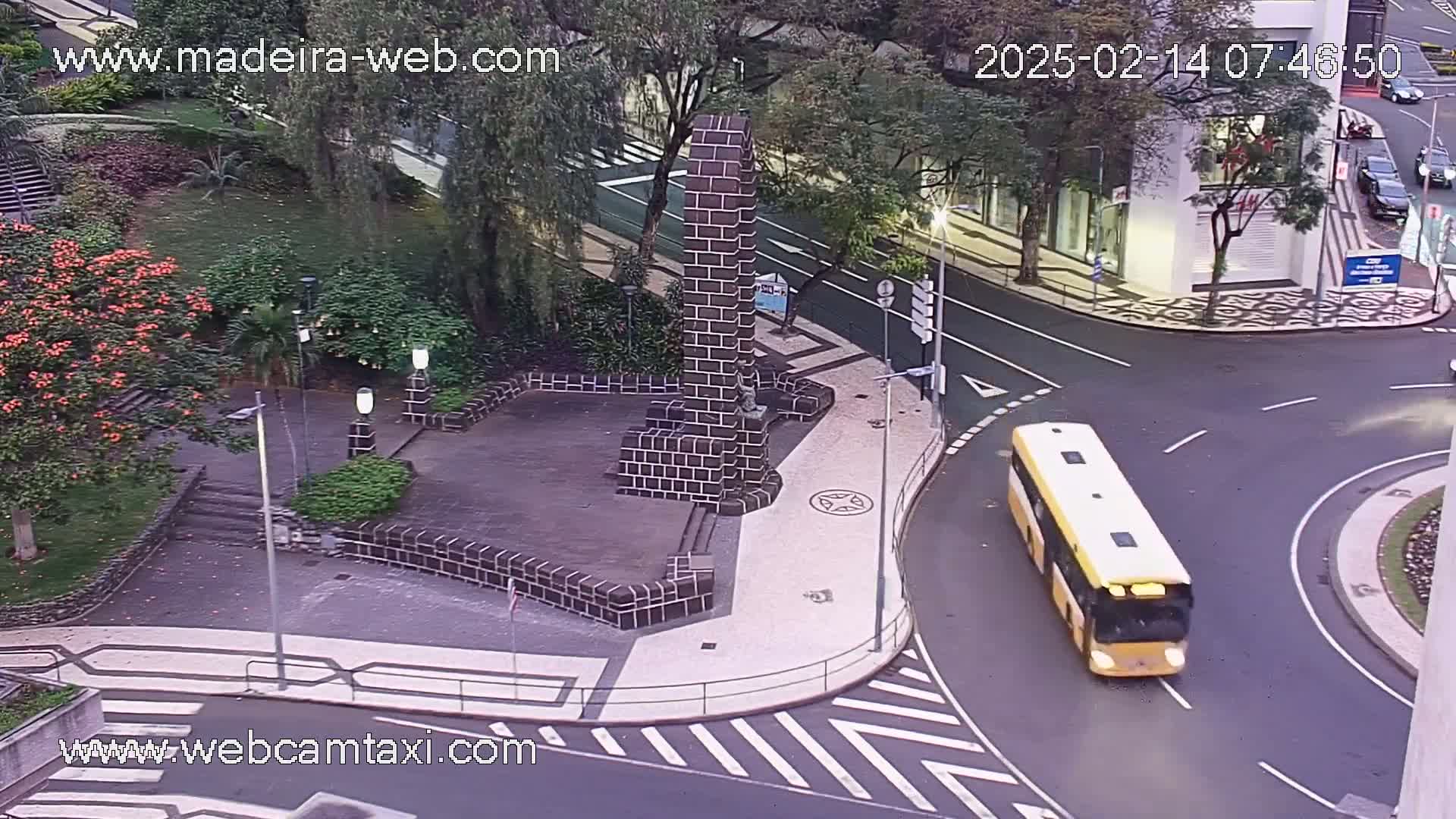 A yellow bus rounds a corner in a city intersection next to a park with a stone monument, under slightly dim daylight conditions.
