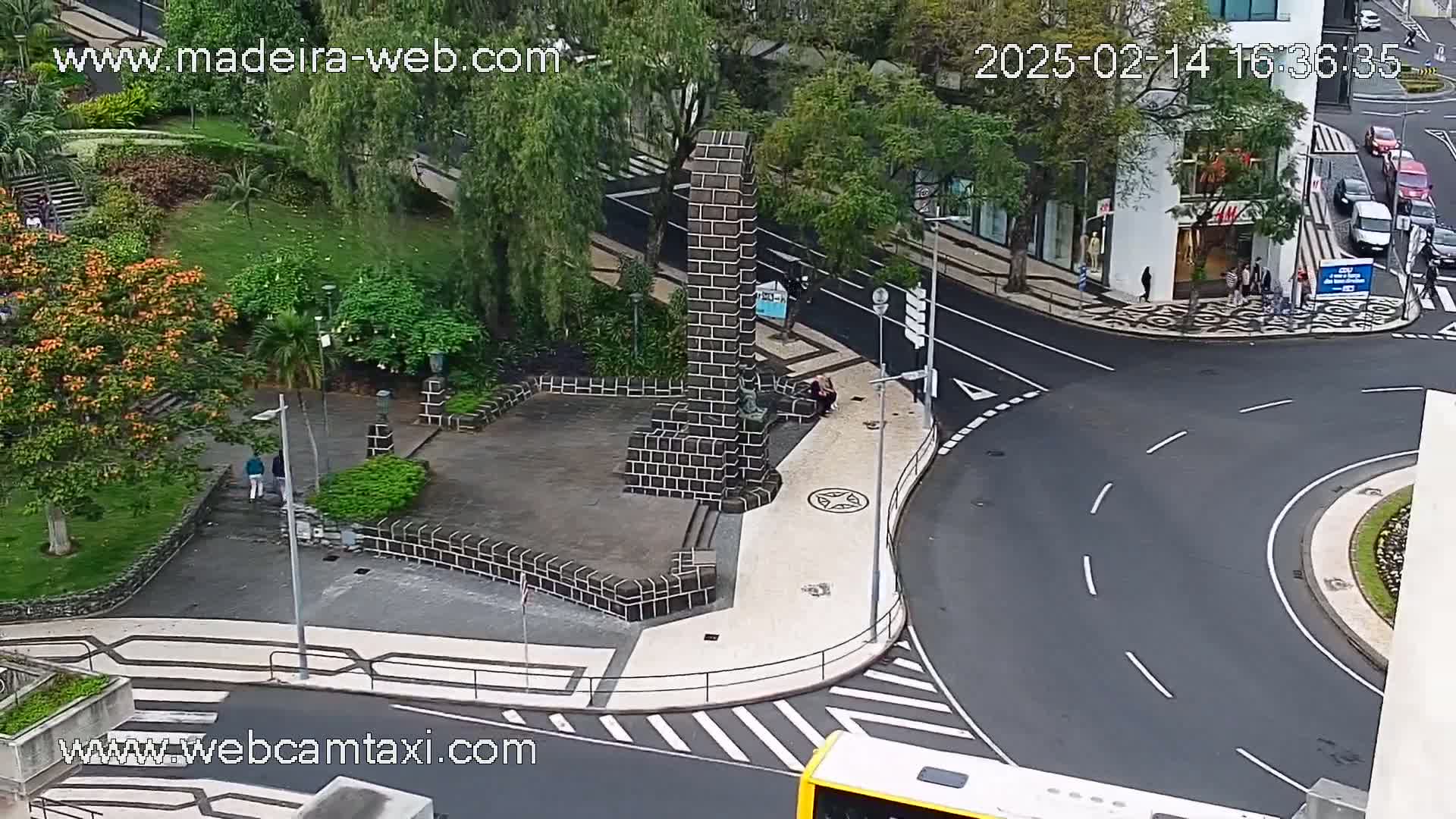 A high-angle view shows a city square with a dark-brick monument, lush green landscaping, and a curving roadway with cars and a bus.