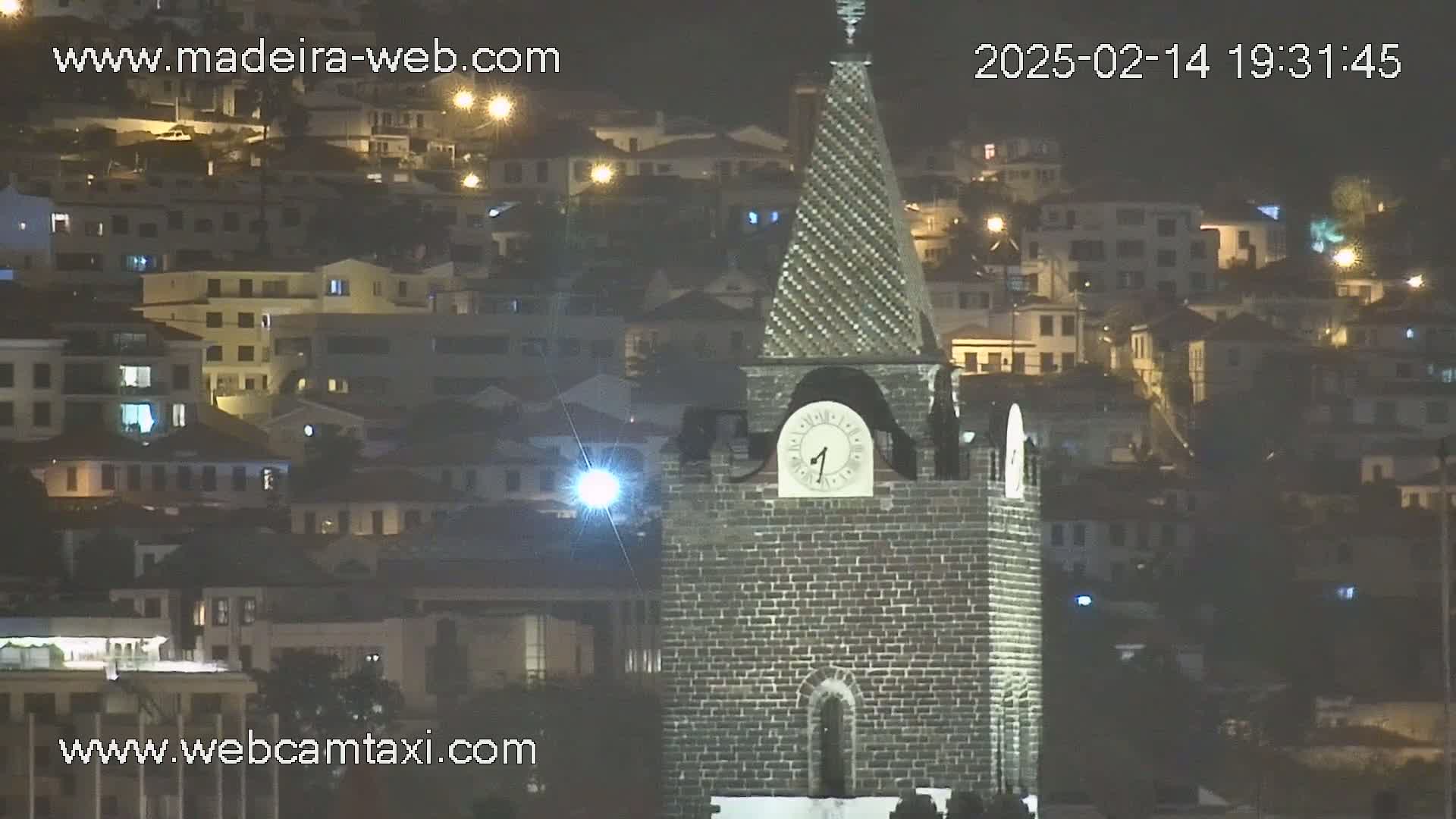 A brick clock tower with a clock showing the time stands prominently in the foreground against a backdrop of a hillside town at night, illuminated by streetlights.