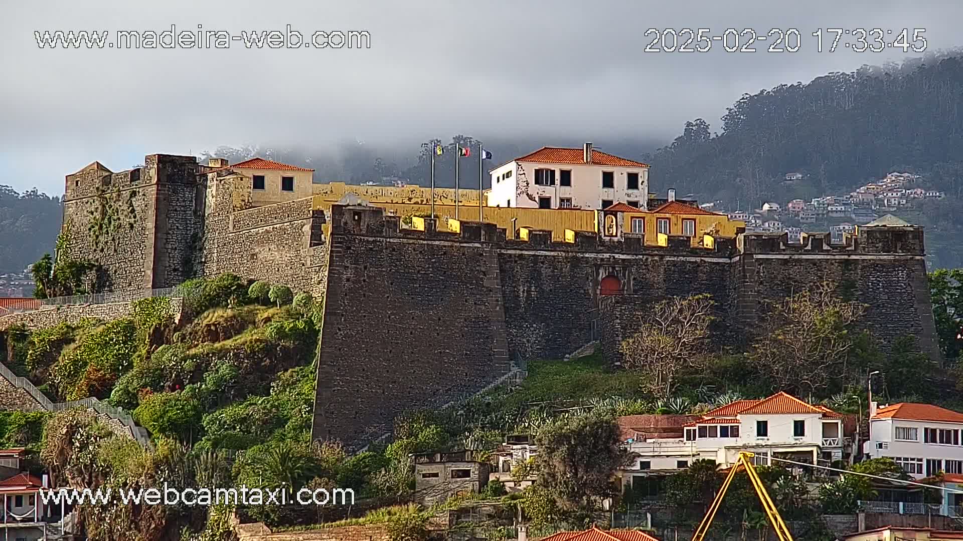 A stone fortress with a few flags on top overlooks a hillside town under a cloudy sky.