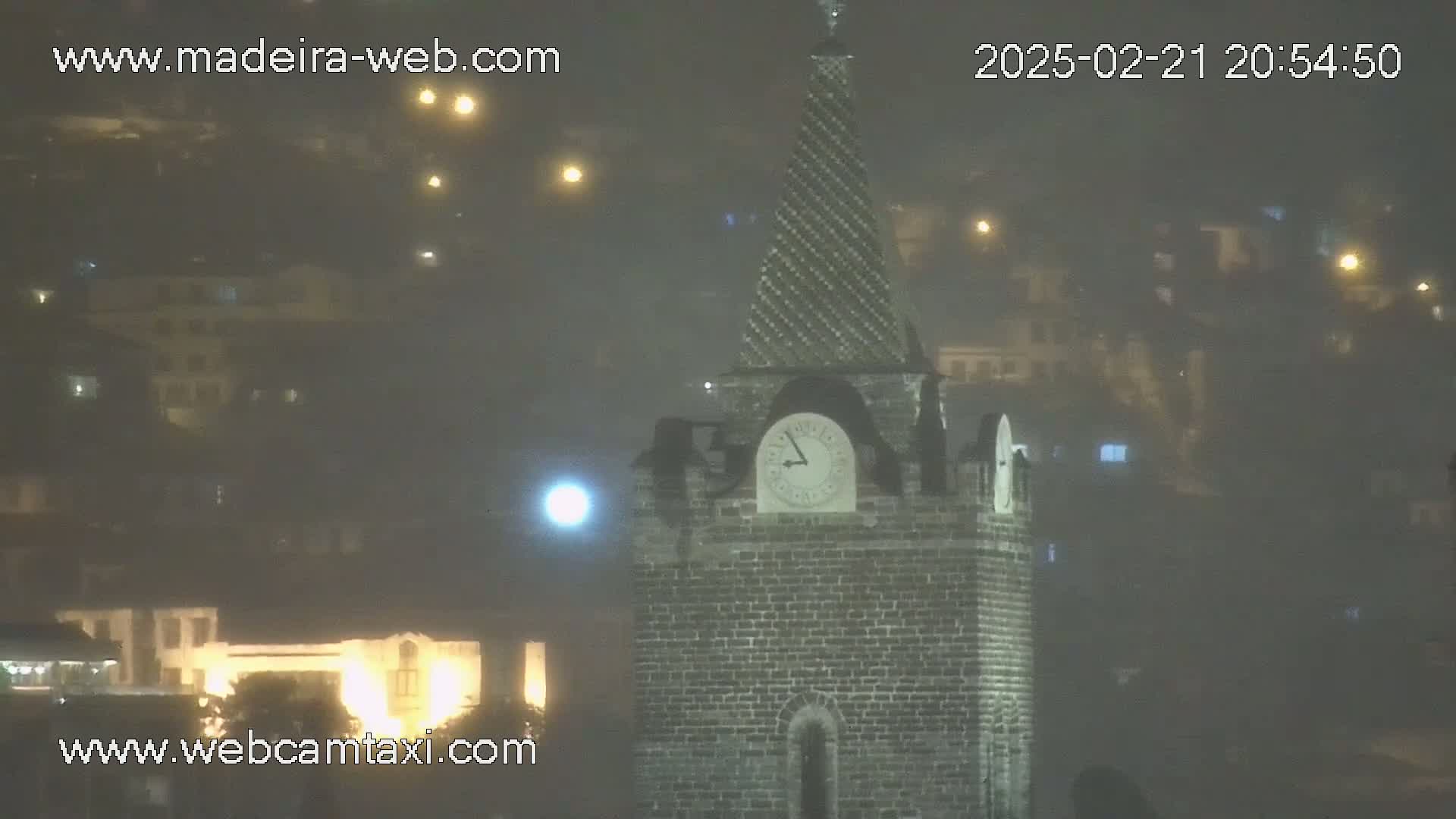 A stone clock tower with a spire stands in a foggy, nighttime cityscape.