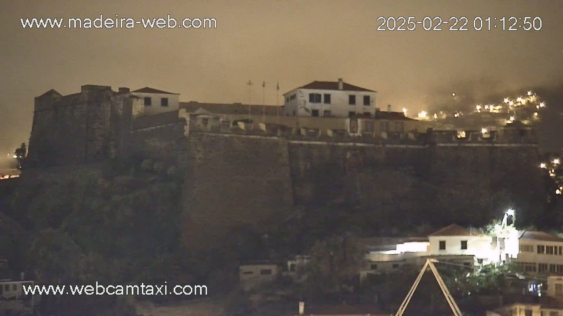 A large, dark stone fortress sits atop a hill at night, under a hazy sky, with other illuminated buildings visible below and in the distance.