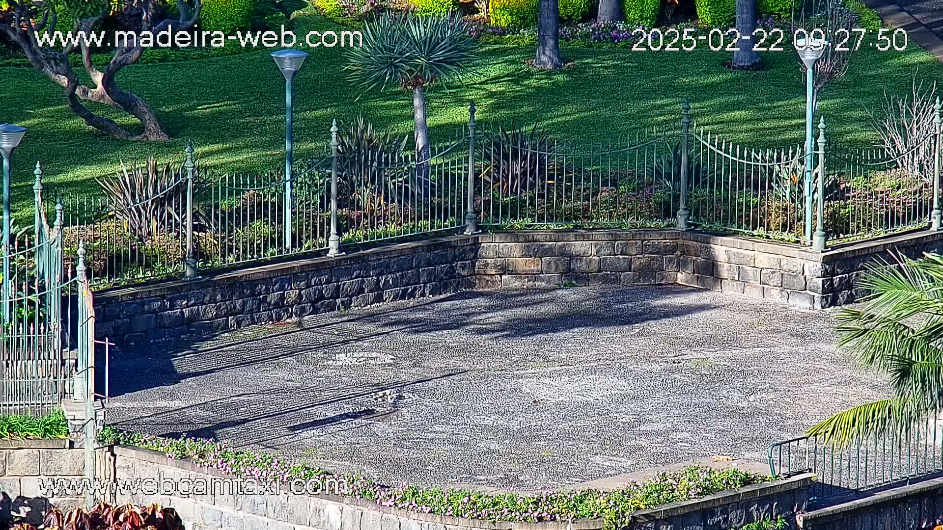 A paved plaza bordered by a low stone wall and an ornate metal fence, surrounded by green lawns, flowering plants, and palm trees on a sunny day.