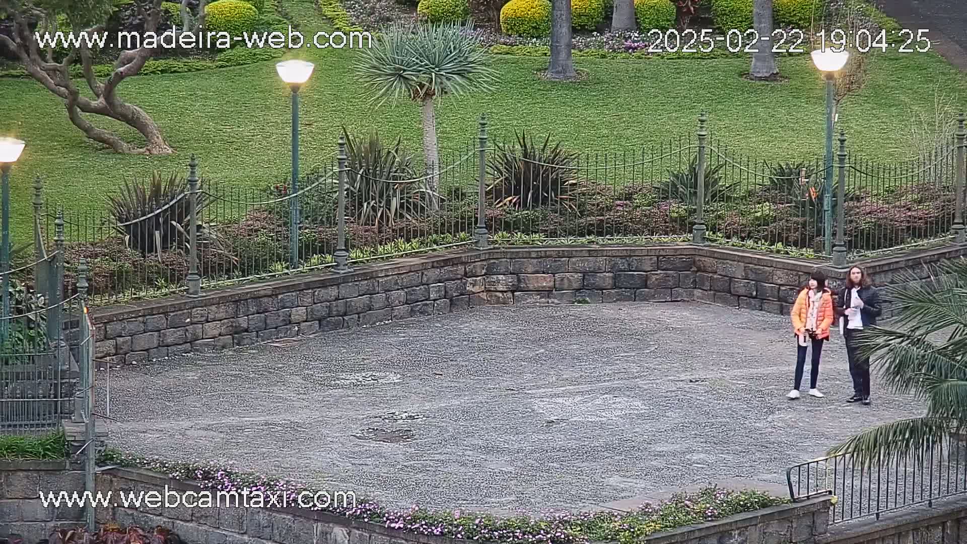 Two people stand on a gray, paved area within a stone-walled garden, under an overcast sky.