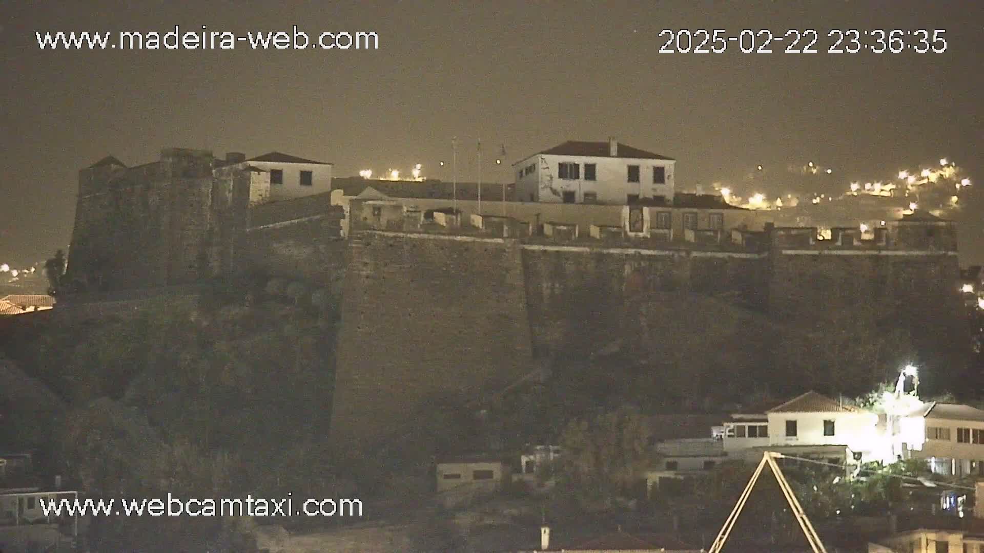 A stone fortress sits atop a hill at night, illuminated by scattered lights, under a dark sky.