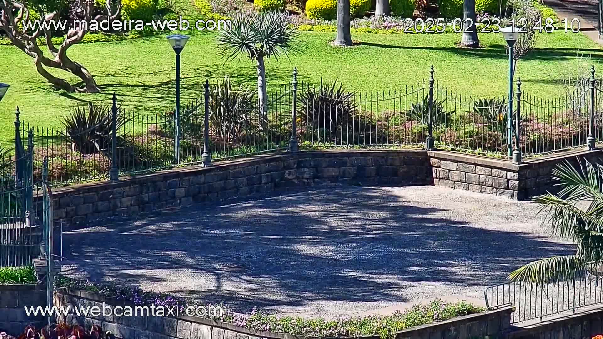A sunny, paved area is enclosed by a low stone wall and wrought-iron fence, surrounded by manicured lawns and flowering plants.