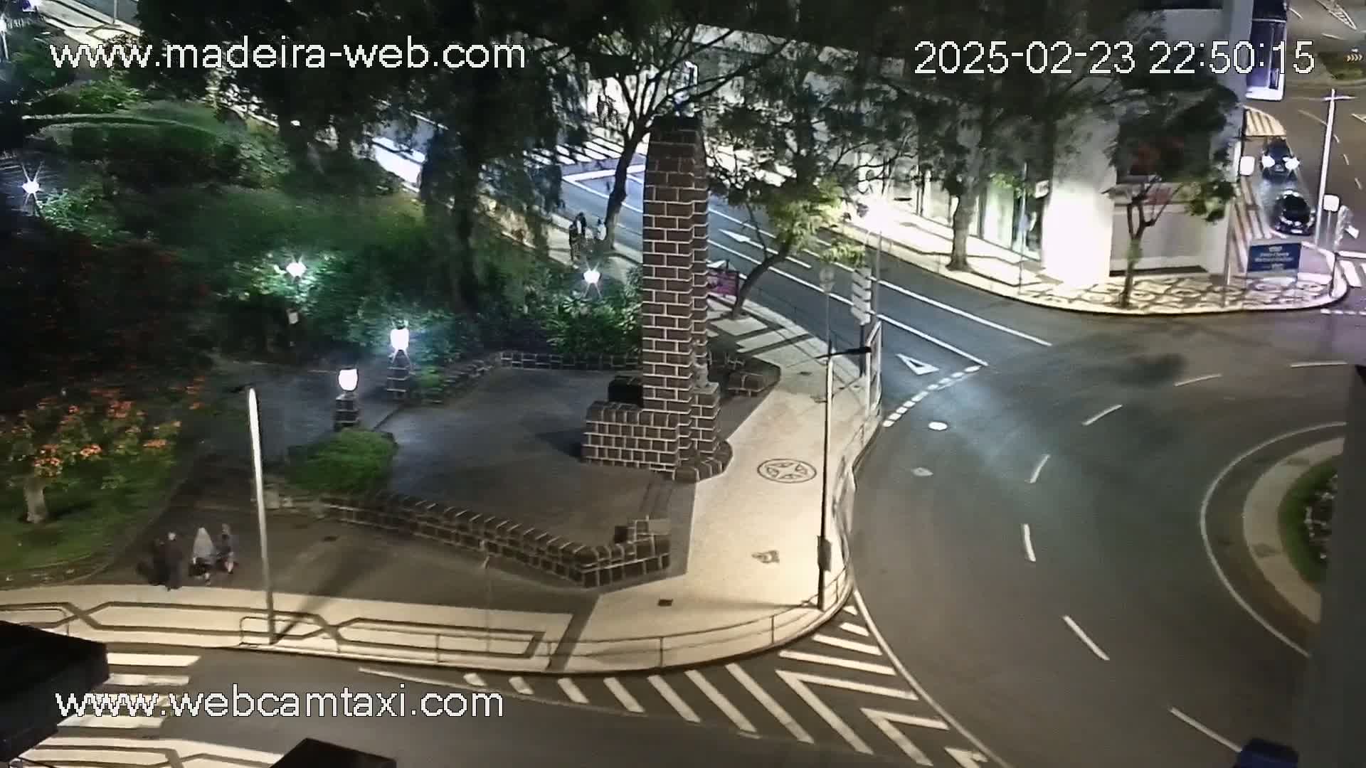 A nighttime view of a paved intersection and a small plaza with a dark brick monument, surrounded by trees and streetlights, with wet roads suggesting recent rain.