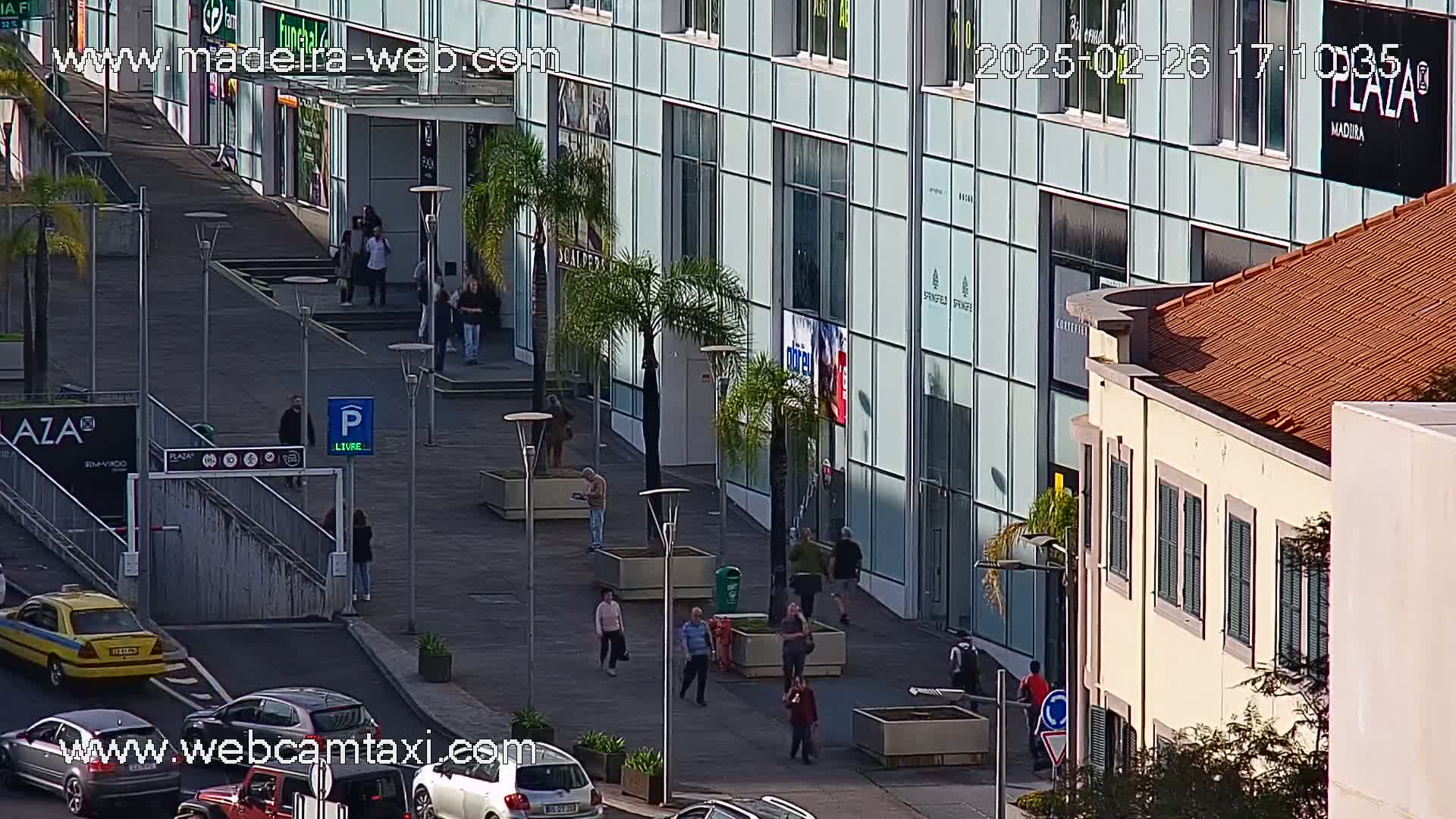 A sunny day reveals a city street scene with pedestrians walking past a modern glass-fronted building and a traditional building with a terracotta roof.