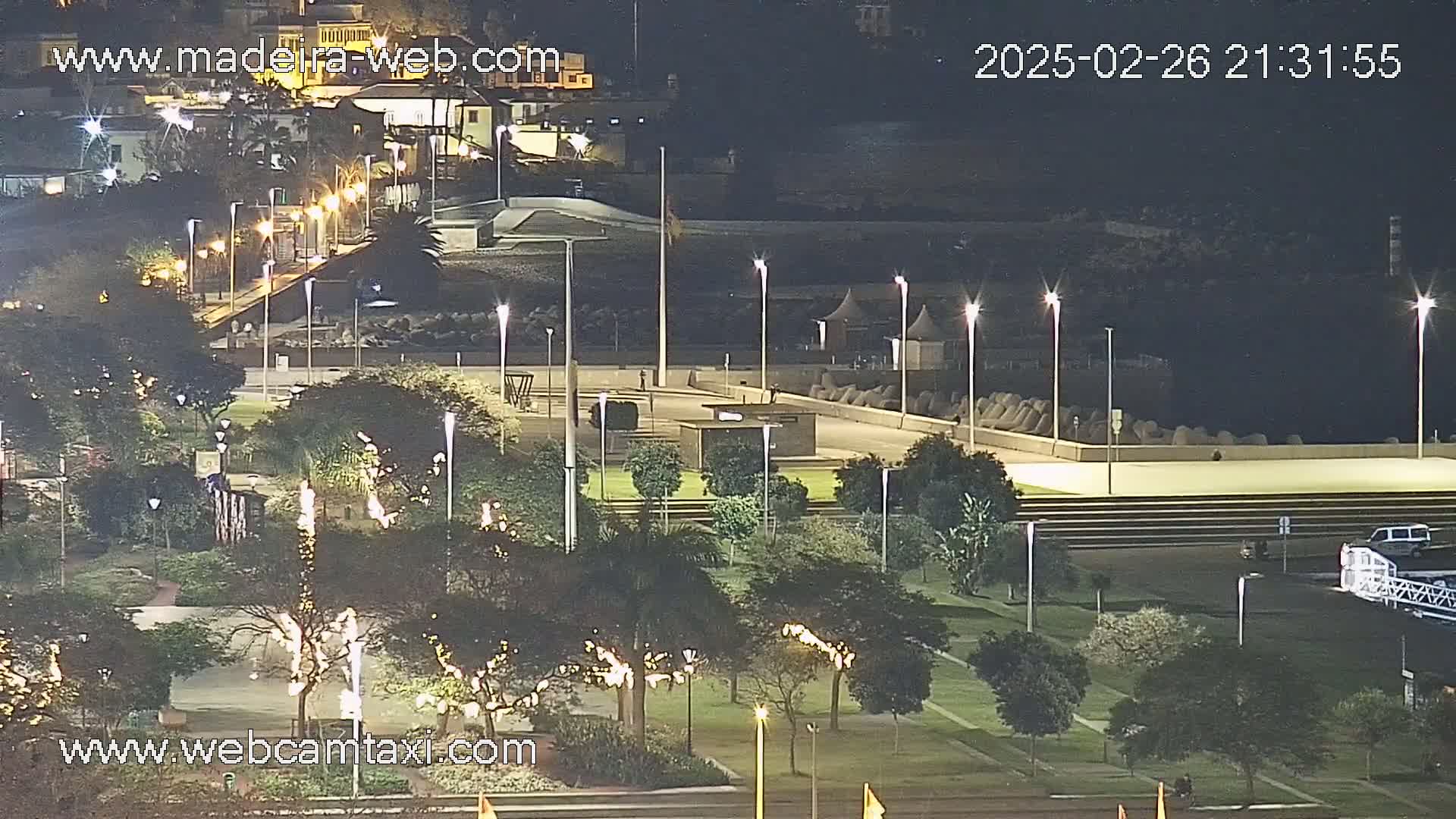 A nighttime view of a park-like area near a waterfront, illuminated by numerous streetlights, with a few buildings visible in the background under a clear sky.