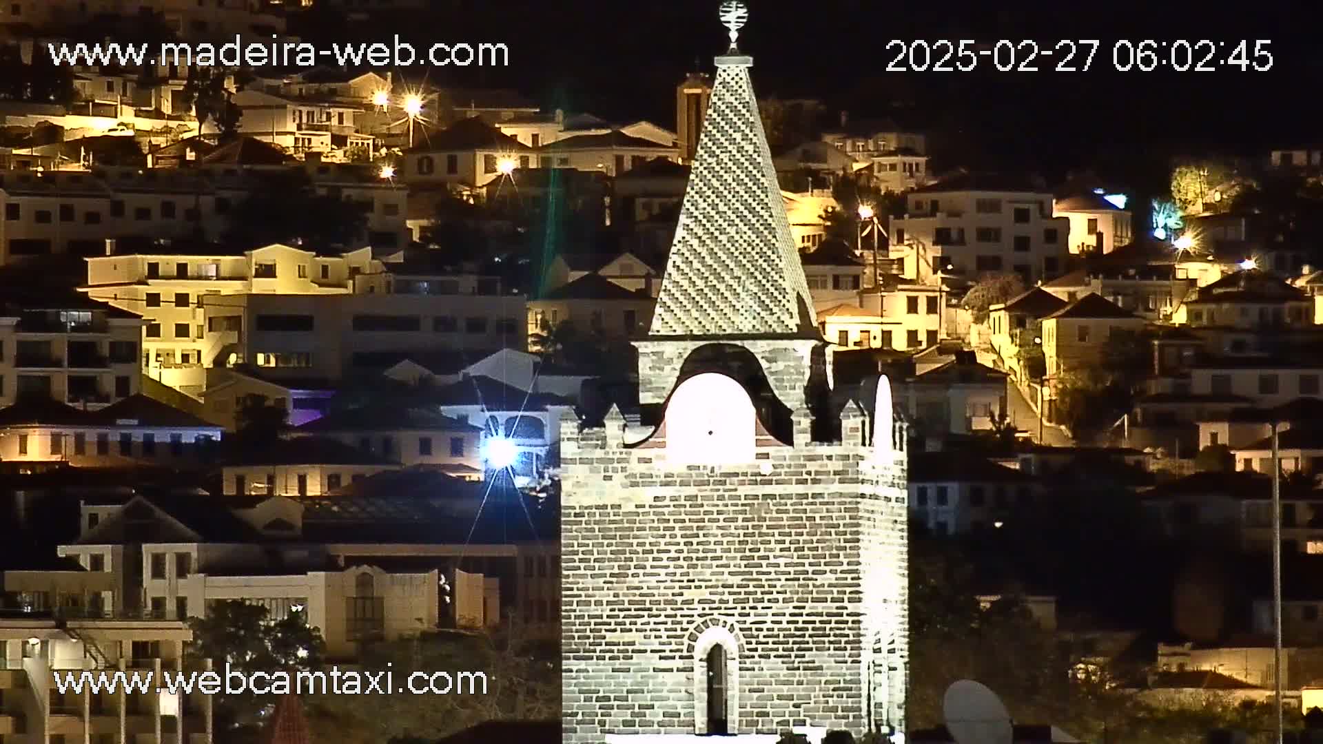A stone tower stands prominently in the foreground, illuminated at night, overlooking a hillside town with numerous buildings glowing with warm interior lights.