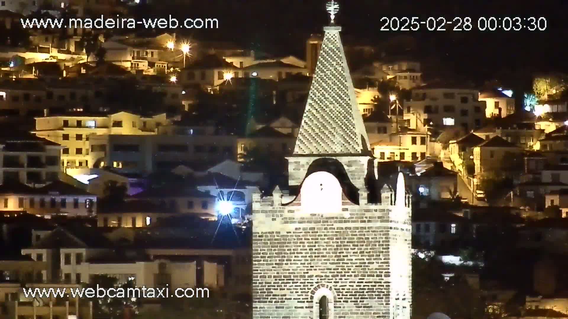 A brick church tower is seen at night, illuminated against a backdrop of a hillside town also lit up.