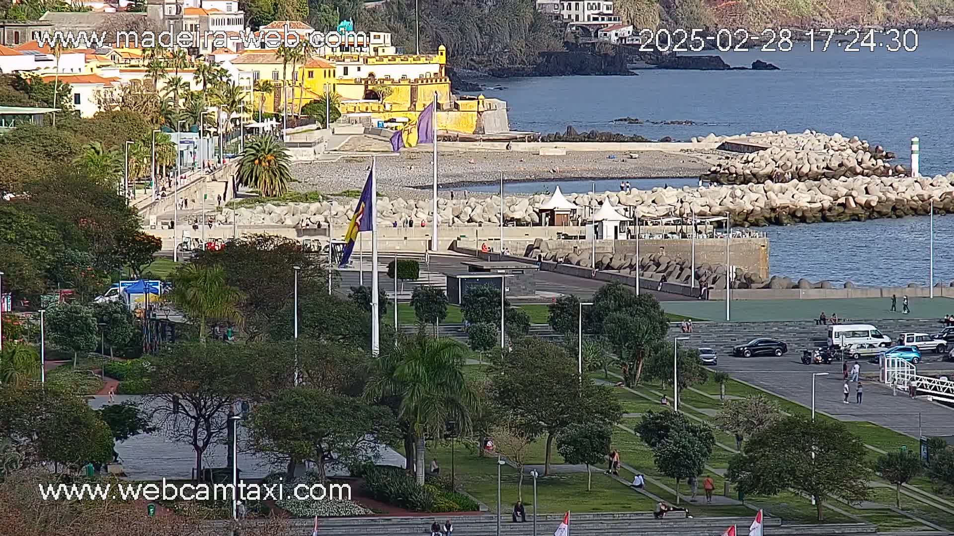 A sunny coastal scene shows a park with people relaxing near a rocky breakwater, a yellow building, and calm ocean water.