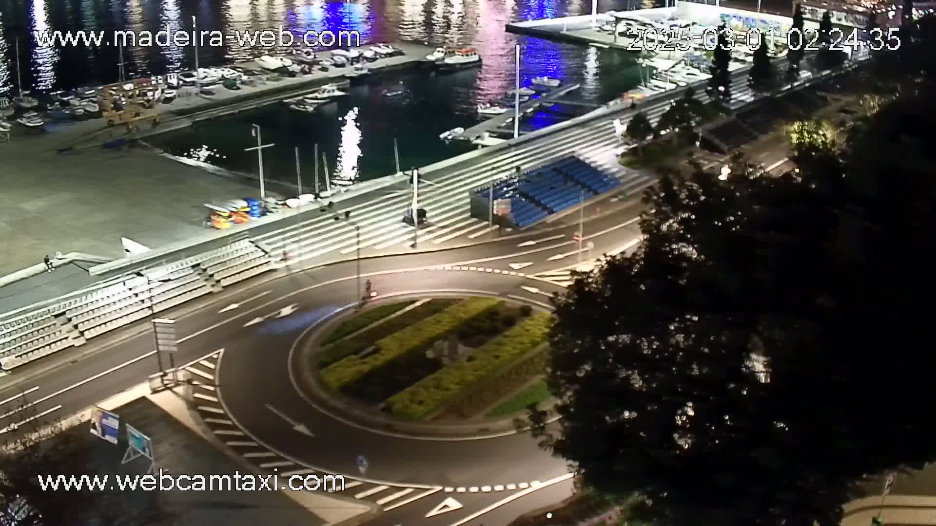 A nighttime aerial view shows a harbor with boats, a roadway with a roundabout, bleachers, and trees.