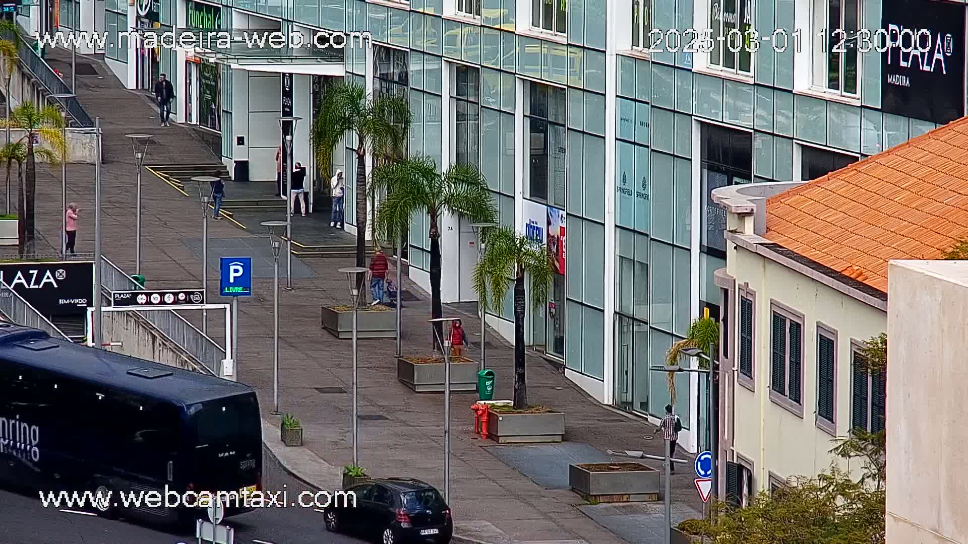 A city sidewalk scene shows a modern glass building and an adjacent older building with an orange tile roof, a few pedestrians, and some parked vehicles.