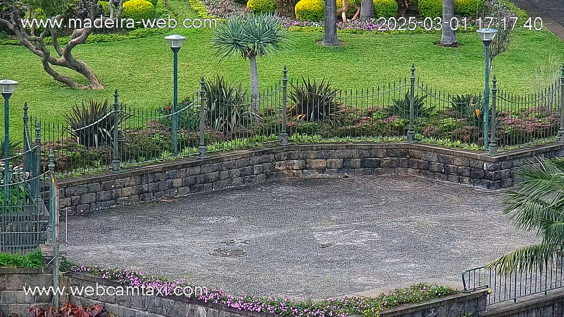 A wet, paved area is surrounded by a stone wall and neatly landscaped gardens with metal fencing and lamps under a lightly overcast sky.