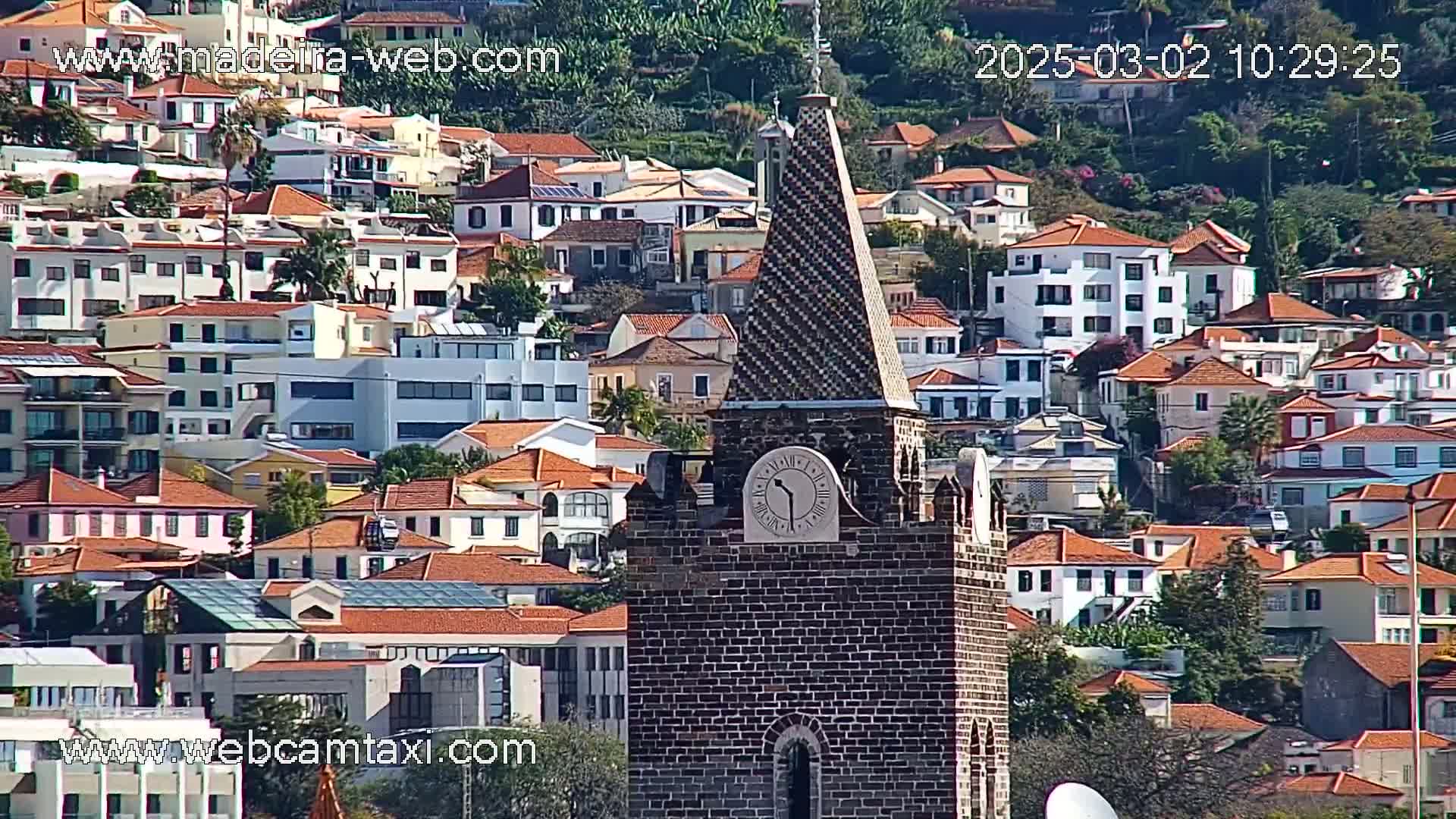 A brick clock tower with a patterned spire stands prominently in front of a hillside densely packed with low-rise buildings, all with red-tiled roofs, under sunny conditions.