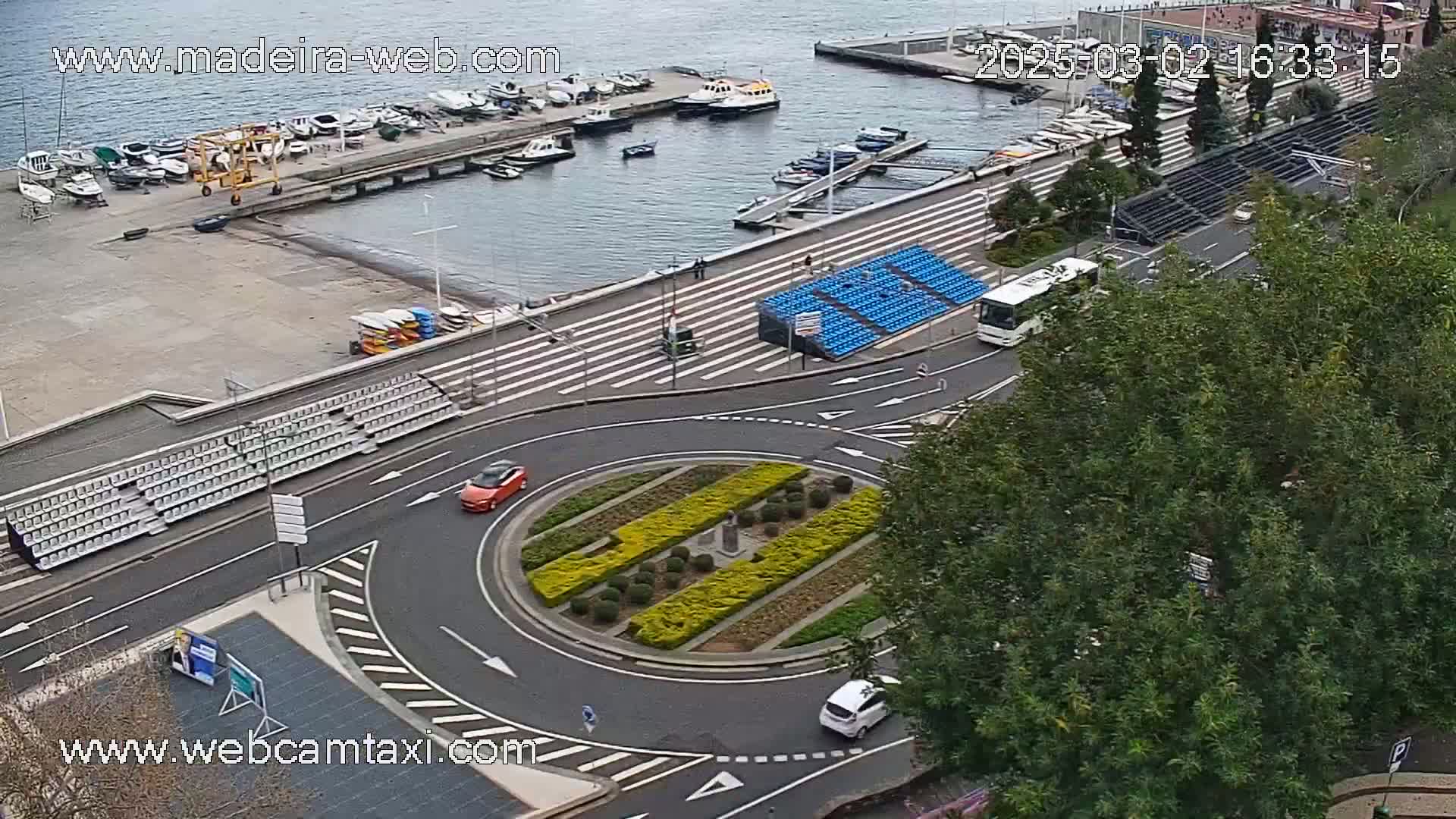 A harbor with numerous boats is visible in the background, while a roundabout in the foreground features two cars and a landscaped center, under an overcast sky.