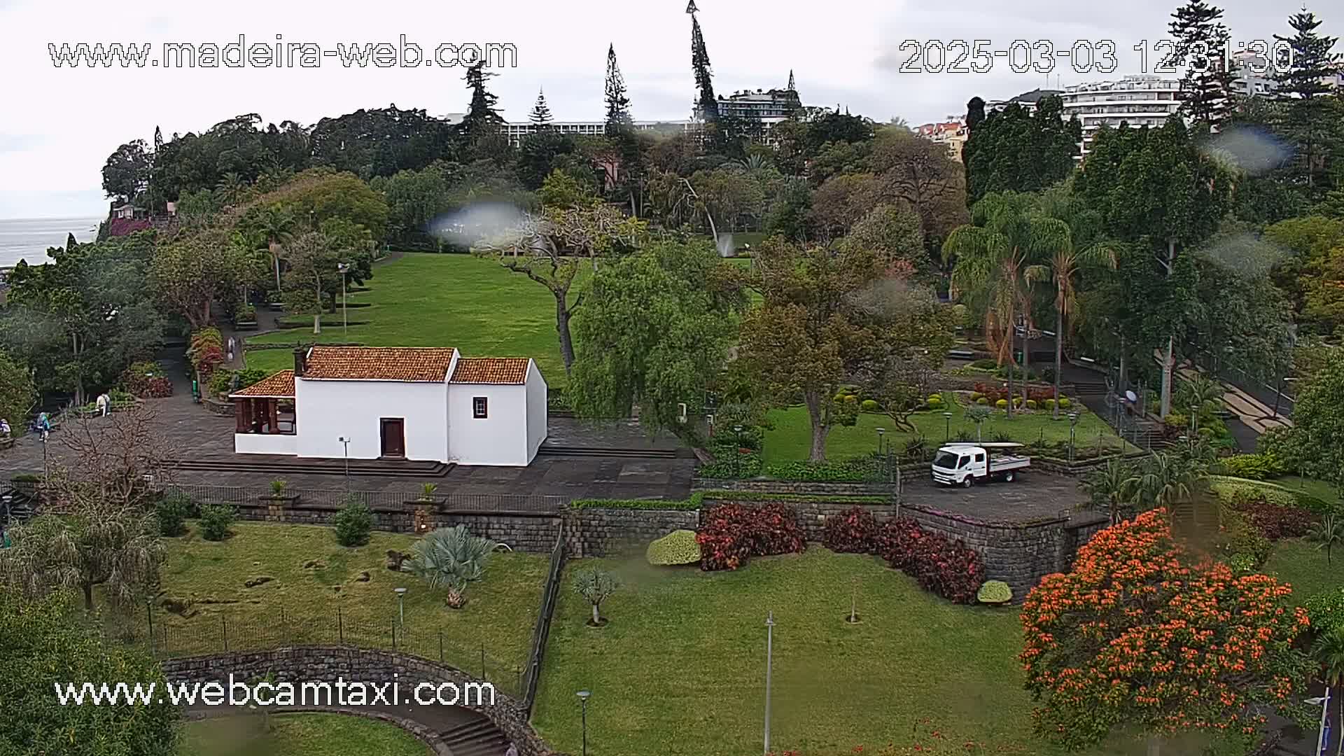 A verdant park with a small white building, lush trees, and a white truck is visible on an overcast day.