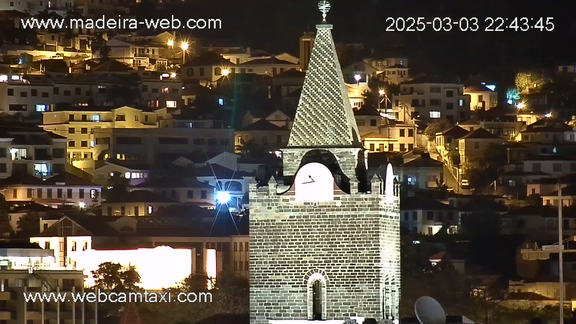 A brick clock tower stands illuminated at night against a backdrop of a hillside town with numerous lit buildings.
