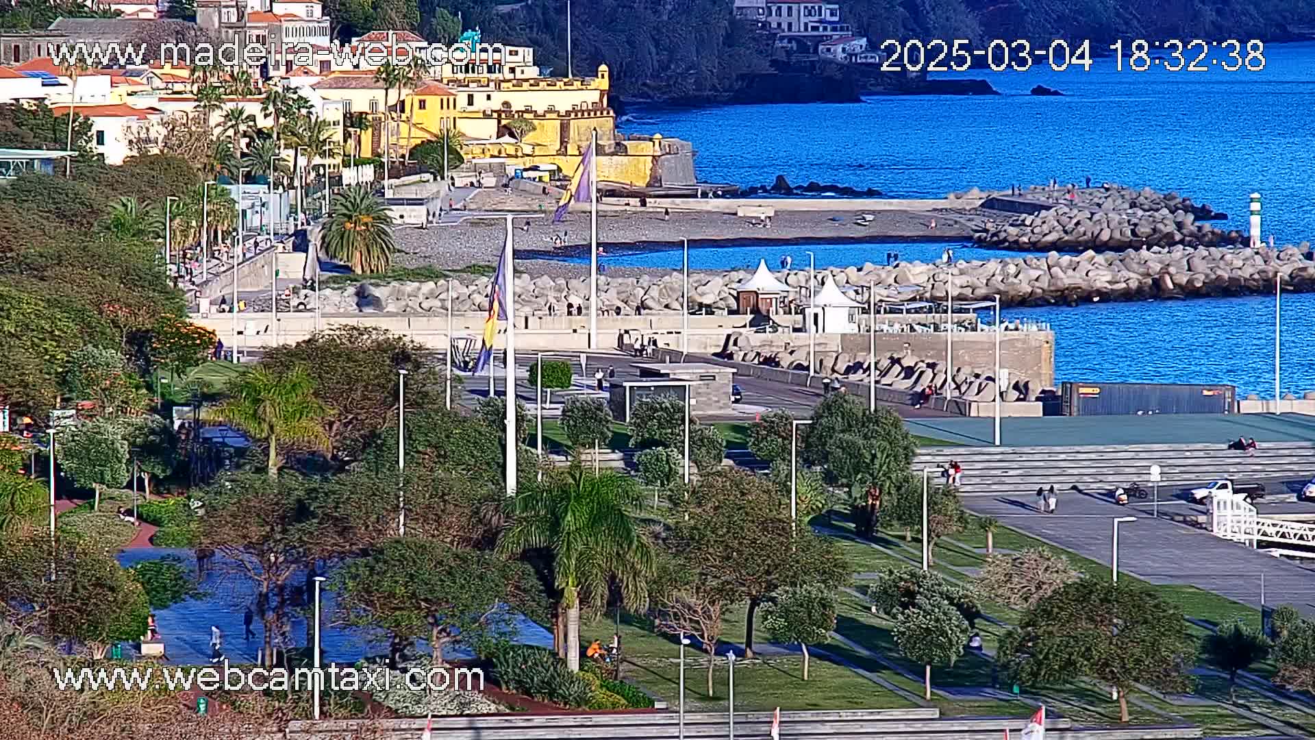 A seaside park with palm trees and paved walkways overlooks a calm, blue ocean dotted with rock breakwaters, near a town of yellow buildings.